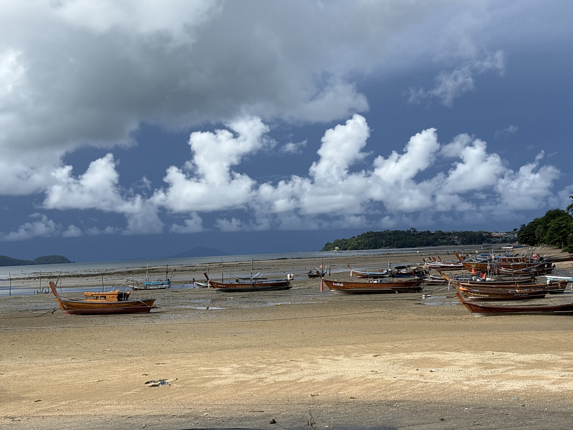 A wide shot shows a beach in Rawai, Thailand, with several wooden longtail boats grounded on the wet sand due to low tide. The boats are of various sizes, some are tied to posts, and others rest directly on the beach. In the background, the calm sea meets a distant, tree-covered island under a dramatic sky filled with dark, heavy clouds interspersed with patches of bright blue and white cumulus clouds. The sandy shore stretches across the foreground and mid-ground, with some debris scattered on the beach. The overall scene depicts a serene but somewhat moody coastal landscape.