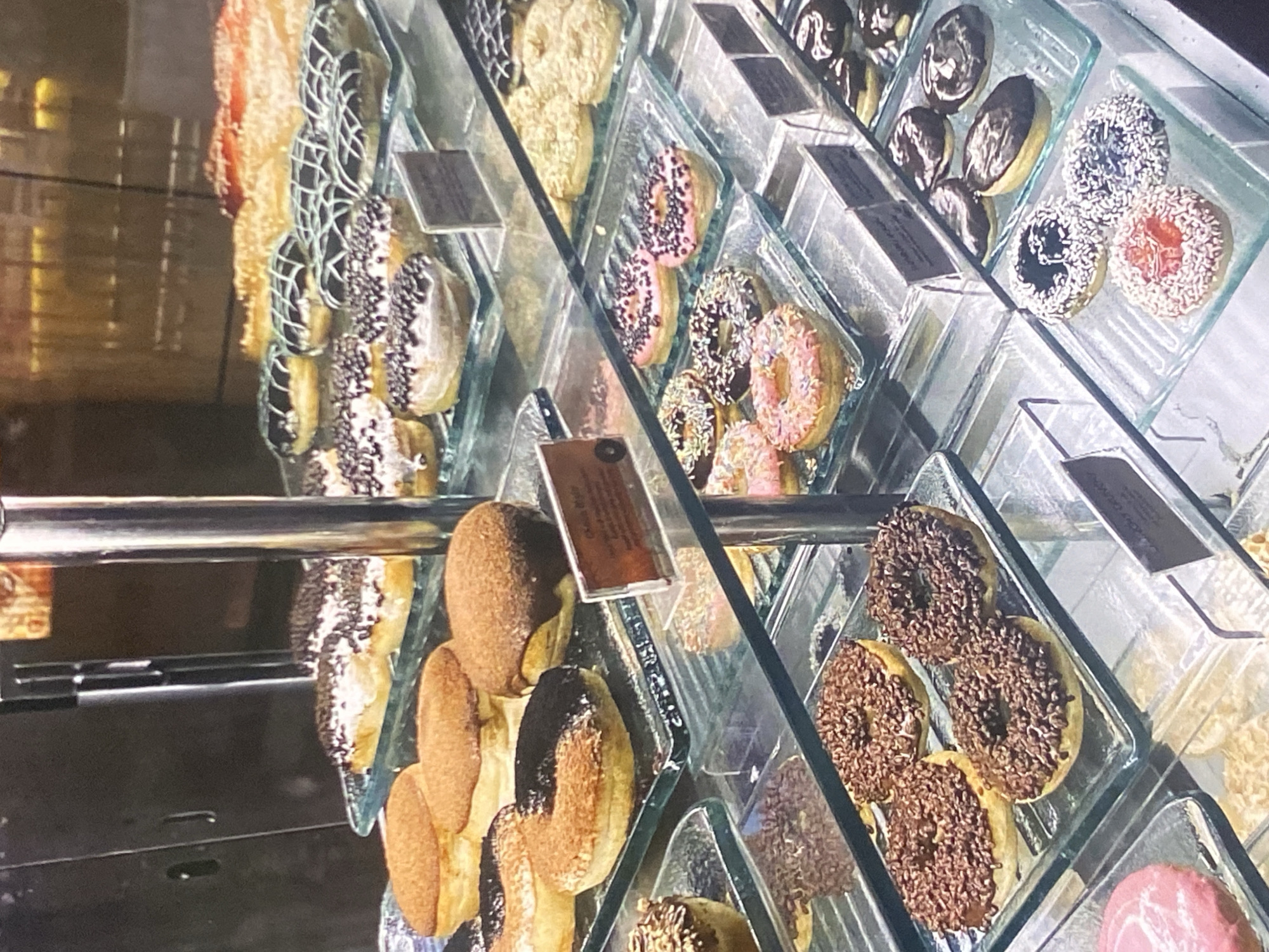 The image shows a display case filled with a variety of donuts. The donuts are arranged on glass shelves, and some have labels with descriptions. The display case is well-lit, highlighting the different textures and toppings of the donuts, which include chocolate glaze, sprinkles, and powdered sugar. The background is blurred but suggests an indoor setting, possibly a bakery or cafe.
