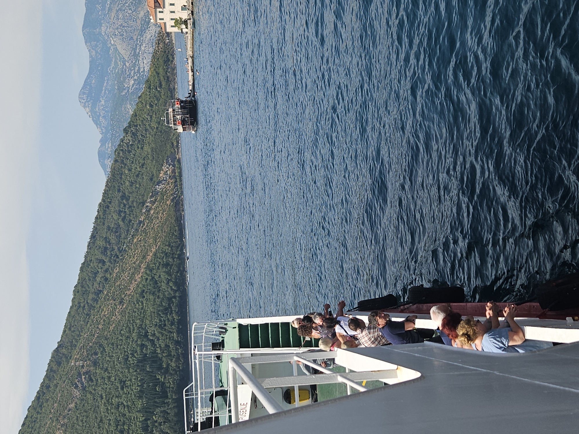 The image shows a ferryboat sailing on the blue sea in Lepetane, Montenegro. A group of passengers are standing on the deck of the ferryboat, looking out at the water. In the background, there are green hills and mountains. The sky is blue and clear. The ferryboat has a white exterior with green accents. One passenger is wearing a blue shirt, another a plaid shirt. The water is calm with small waves. The scene suggests a scenic boat ride on a clear day.
