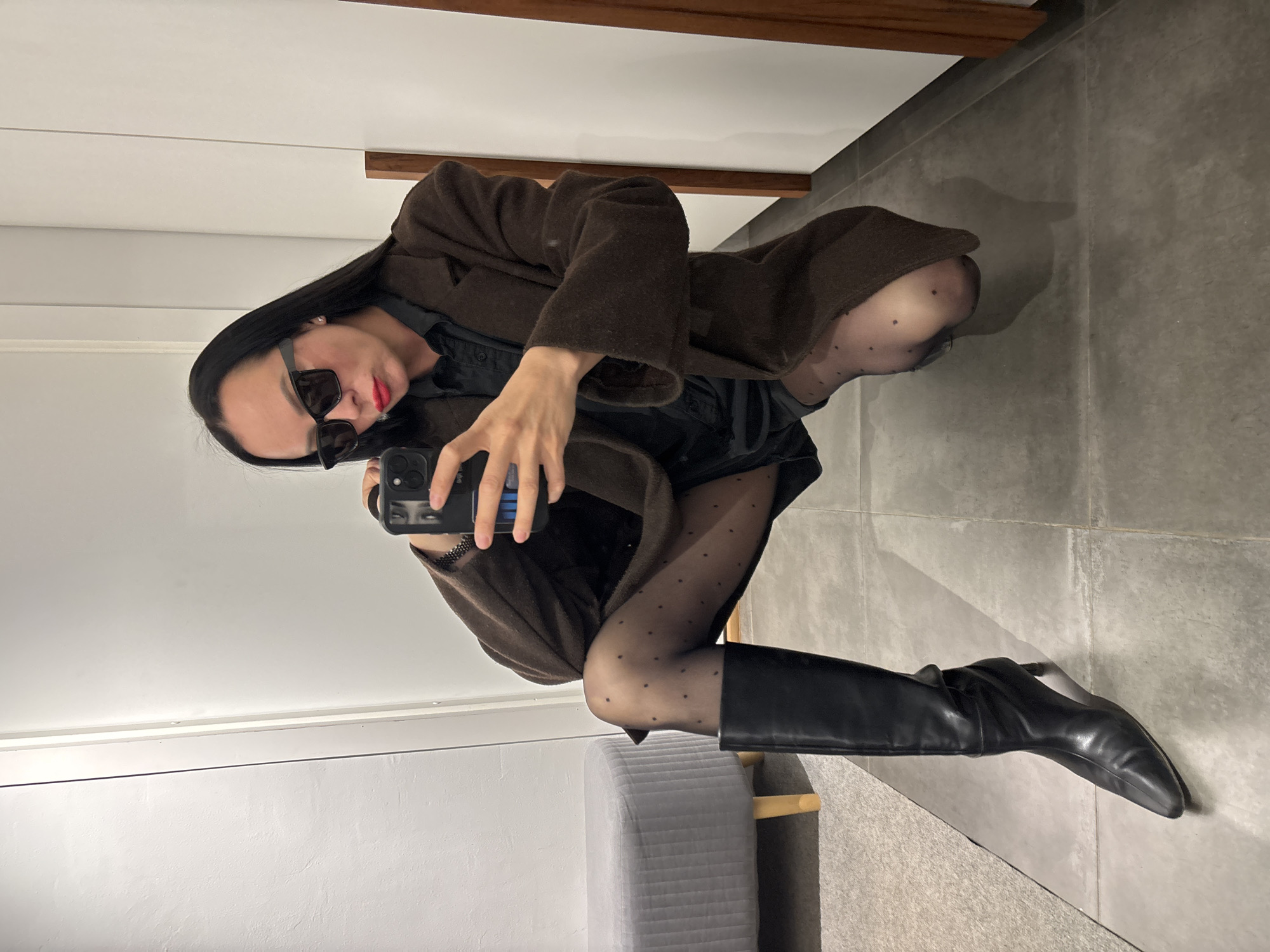 The image displays a full-body mirror selfie taken by a person indoors. The subject, likely female, is captured in a crouching or squatting pose, holding a black smartphone to take the photo. The image itself is rotated 90 degrees clockwise.
The person has long, straight dark hair and wears dark, rectangular sunglasses that obscure their eyes, along with vibrant red lipstick. They are dressed in a dark brown, long-sleeved coat over a dark-colored top or dress. Below the coat, they wear sheer black tights adorned with small, evenly spaced polka dots. The footwear consists of sleek black knee-high boots with pointed toes and heels.
The indoor setting reflected in the mirror features a grey tiled floor and a clean white wall. A horizontal brown wooden beam or ledge is visible at the top of the reflection. In the lower left, a section of grey upholstered furniture, possibly a bench or chair back, is present. The lighting is even, suggesting an indoor environment. There is no discernible text visible in the image. The location of the photograph is Chaikovsky, Russia.