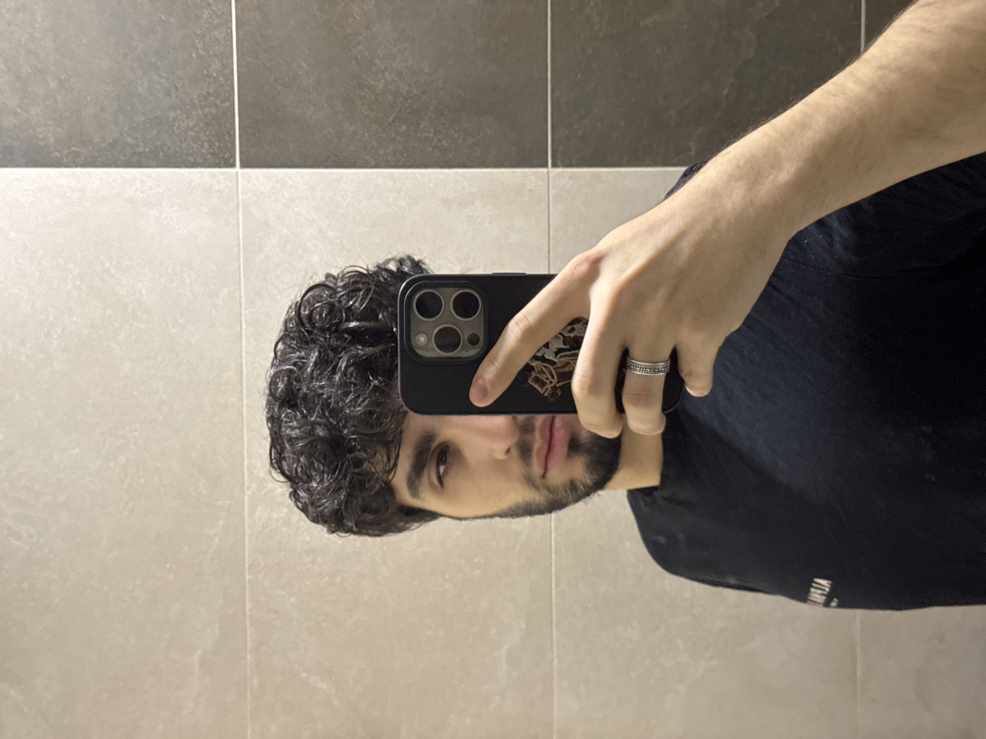 The image depicts a male individual taking a mirror selfie in an interior location within Yerevan, Armenia, likely a bathroom or washroom. The subject has dark, curly hair which appears wet, and a beard. He is wearing a dark-colored t-shirt with the mirrored text "АРМЕНІА" (ARMENIA) visible on his left chest/shoulder area. His right hand holds a dark-cased smartphone with a prominent multi-lens camera system visible on its back, obscuring the right portion of his face. A silver-colored ring with textured detailing is worn on the ring finger of his right hand. The background is composed of tiled walls; light beige-toned rectangular tiles are visible on the left and upper right, while darker grey-brown tiles are present on the lower right, arranged with visible grout lines.
