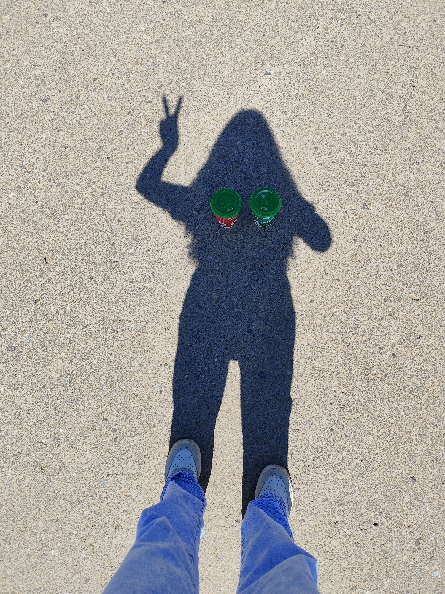 A top-down view captures a person's shadow cast on light gray, speckled concrete or asphalt ground in Фоки, Russia. The shadow, appearing to be that of a woman, shows her right arm raised with her hand forming a "peace" sign gesture. Two small, lidded beverage cups are placed on the ground within the shadow, approximately where the person's torso would be. Both cups have bright green lids. The body of the left cup is predominantly red, while the right cup's body is green. Although difficult to read definitively, partial text on the cups includes what looks like "Premium" and possibly "Fresh," but a specific brand name is not clearly discernible.
Below the shadow, the person's feet are visible, wearing light-colored sneakers with visible blue shoelaces, paired with blue denim jeans. The sharp, distinct edges of the shadow indicate bright sunlight, suggesting a clear, sunny day, likely mid-morning to early afternoon. The setting is an outdoor paved area. The overall impression conveyed is casual and positive, emphasized by the peace sign.