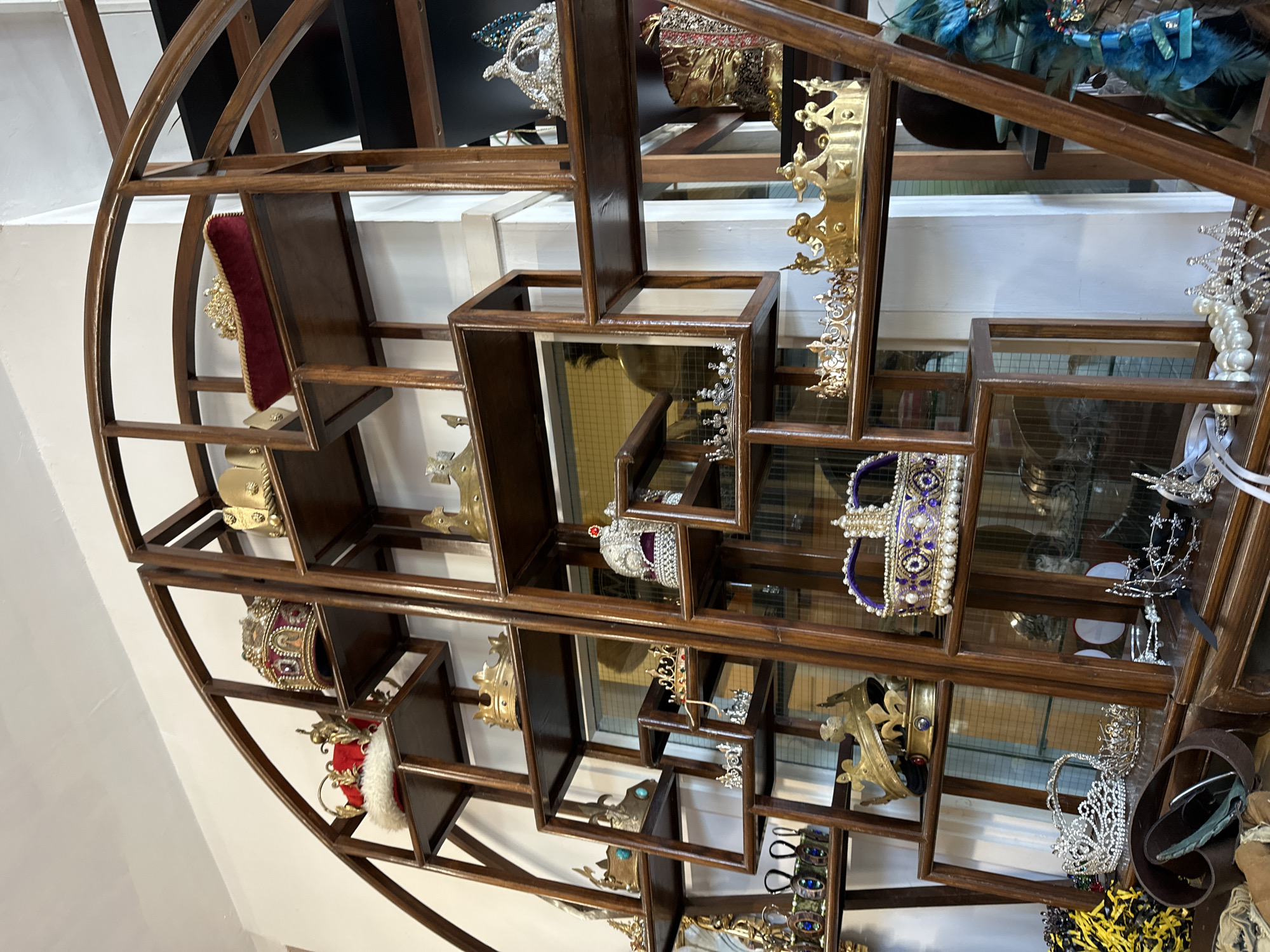 A tilted close-up shot shows a round, wooden display shelf filled with various decorative crowns. The shelves themselves are of varying sizes and shapes, creating an irregular grid pattern within the circular frame. The crowns vary in style and material, some being gold, silver, or jewel-encrusted. Reflections are visible on the mirrored shelves, adding depth and visual complexity to the scene. The backdrop is a mix of light walls and dimly lit spaces, but the precise location in London is unclear from the given visual information.