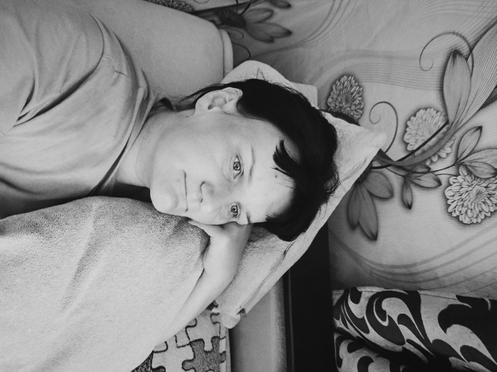A woman lies on a patterned couch. The image is black and white. The woman is resting her head on a pillow and her hand is near her face. The background features floral patterns. The location is unknown and is not in Фоки, Russia.