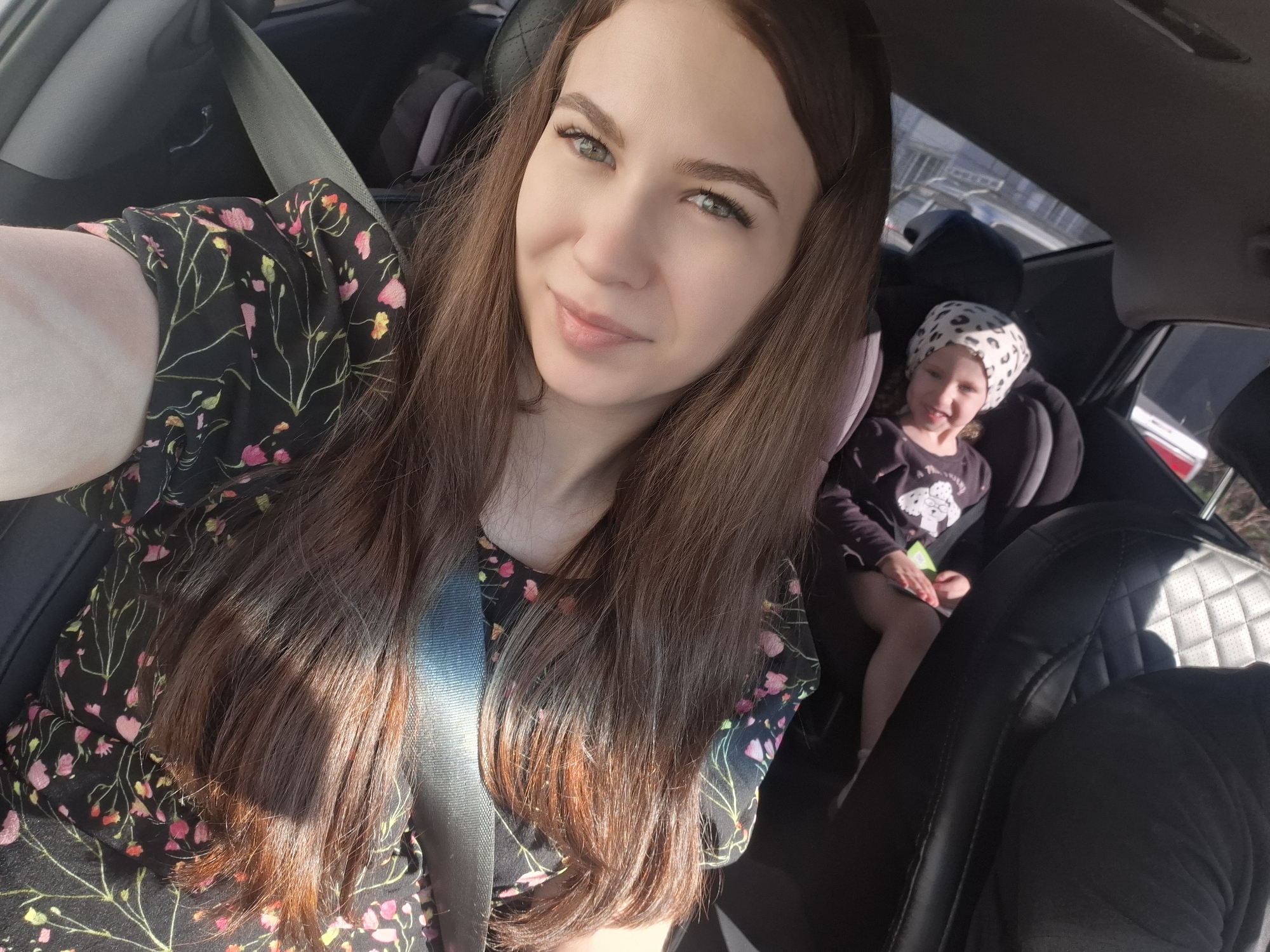 The image shows a woman taking a selfie in the driver's seat of a car. She has long brown hair, green eyes, and is wearing a black floral-patterned dress with a seatbelt fastened across her chest. In the back seat, a young child wearing a spotted headscarf is seated in a car seat. The location is likely Chaikovsky, Russia. The environment appears to be a car interior with glimpses of the outside through the windows.