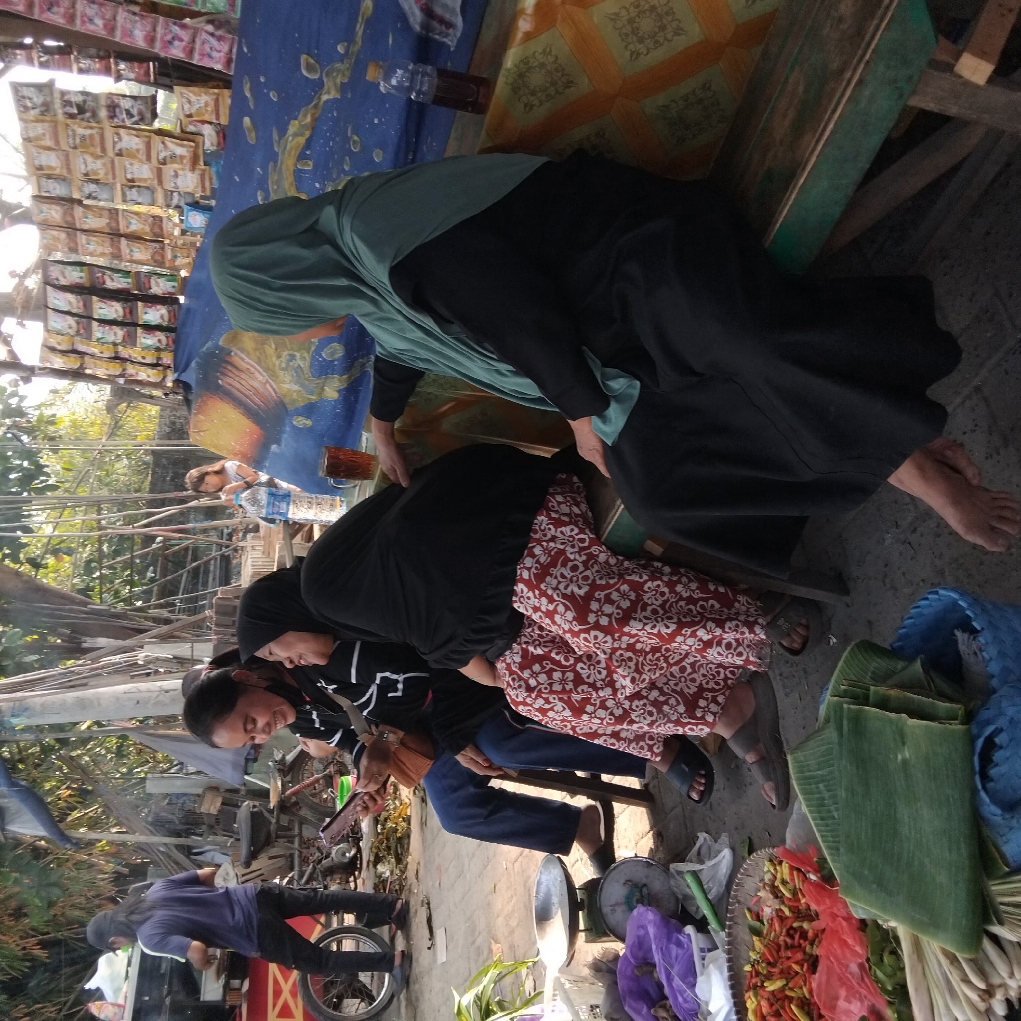 In Banjarbaru, Indonesia, at an outdoor market, several individuals are gathered. One person, wearing a blue shirt and a cap, stands near a food stall. Three women are seated on a wooden bench. The woman on the left, dressed in a black and white striped shirt and blue jeans, is looking at her phone. Next to her, a woman in a black hijab and a red and white patterned skirt is laughing. The third woman, on the right, wears a green hijab and a black outfit and appears to be talking to the woman next to her. In the foreground, a weighing scale, a basket filled with chilies, and banana leaves are visible. The background shows a stall with various packaged goods displayed.