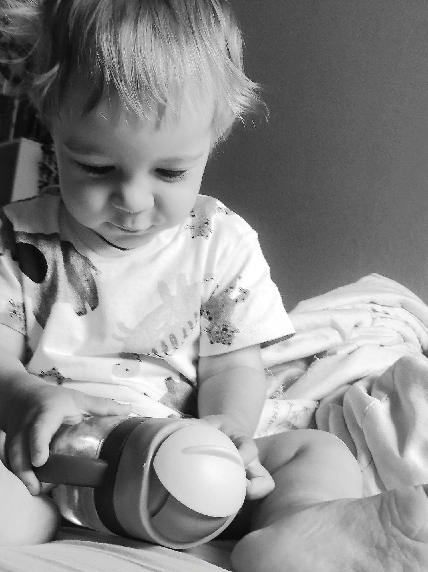 This black and white image captures a young child, likely a toddler, seated in an indoor setting, possibly on a bed or sofa. The child is the central subject, with their head tilted down and eyes focused on an object held in their hands. They have light-colored, slightly tousled hair and soft facial features, conveying a sense of quiet concentration or contemplation.
The child is wearing a light-colored t-shirt with repeating patterns of cartoon animals, possibly bears or cats, visible on the sleeves and torso. Their bare arms and legs are visible, resting on soft, light-colored bedding or blankets. In their hands, the child holds a clear water bottle or sippy cup, featuring a dark-colored handle or grip and a prominent light-colored, rounded spout or lid. They appear to be grasping the bottle firmly, either preparing to drink or engaged in play.
To the right and behind the child, folded or bunched light-colored fabrics, consistent with bedding or blankets, are present. Faint text, partially visible on one of these fabrics, appears to spell out "MAM". The background is largely dark and out of focus, emphasizing the child and their immediate activity. The image evokes a calm and intimate moment. The location context for this scene is Chaikovsky, Russia.