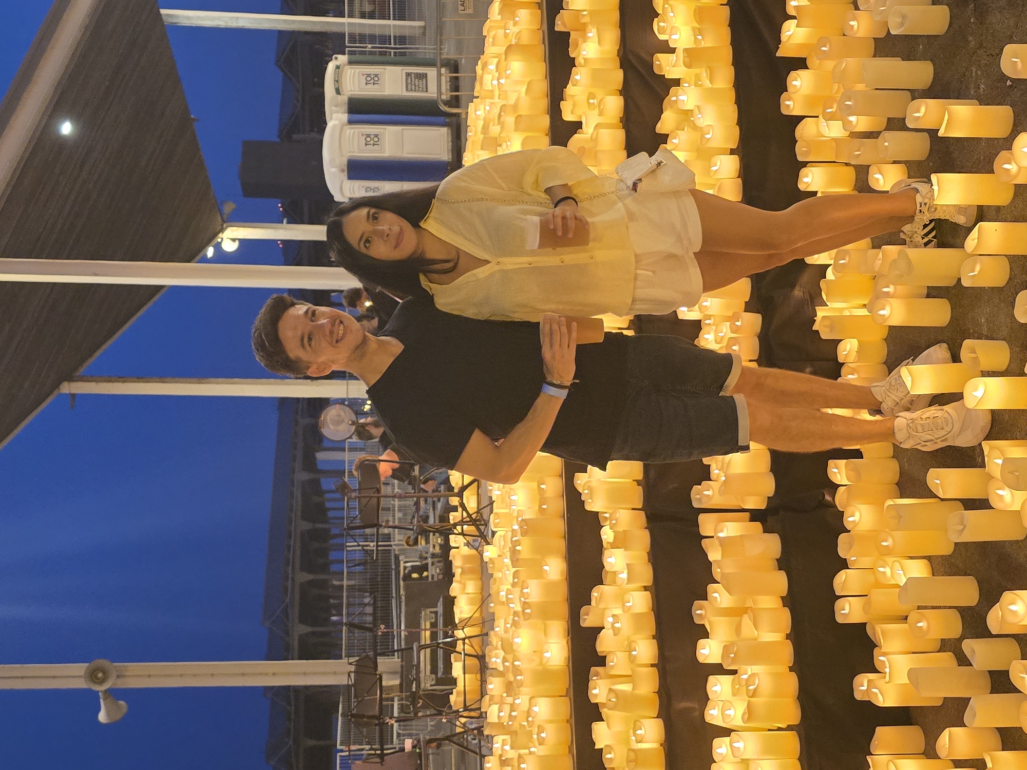 The image depicts a couple standing in front of rows of lit candles. The man, wearing a black t-shirt and dark shorts, holds a plastic cup. The woman, in a yellow button-up shirt and white shorts, also holds a plastic cup and wears a white purse. They are both looking towards the camera and smiling. The background shows a large outdoor space with a covered structure, some portable toilets, and stage equipment. The setting appears to be at dusk or night, with the candles providing the primary light source. The overall atmosphere is festive and warm. The location of the scene is the Parc del Fòrum in Sant Adrià de Besòs, Spain, which is a large public space known for hosting events and concerts.