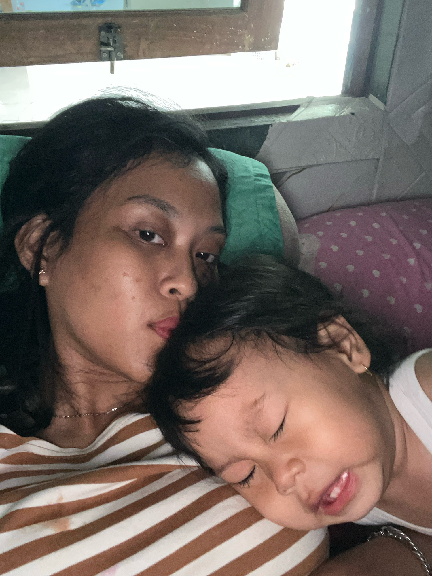 A woman and a child are lying down, likely in a bedroom in Kecamatan Pati, Indonesia. The woman, with dark hair and brown eyes, is looking towards the camera with a relaxed expression. The child, also with dark hair, is sleeping with their head resting on the woman's shoulder, their eyes closed and mouth slightly open. They are both wearing casual clothing. The background shows a window with a view of the outdoors and a patterned pillow. The overall scene conveys a sense of intimacy and rest.