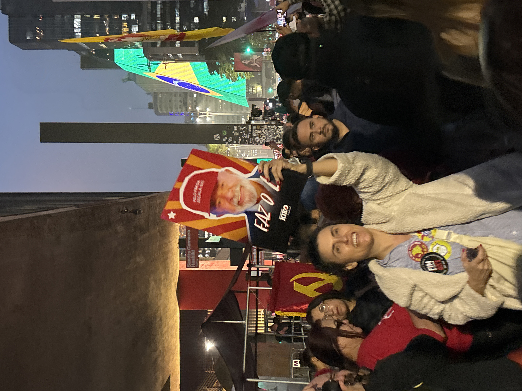 A nighttime photograph captures a political gathering in São Paulo, Brazil, identifiable by the prominently displayed Brazilian flag illuminated on a building. A crowd of people is visible; a woman in the foreground smiles while wearing a light blue shirt with several buttons and a white cardigan. She holds a mobile phone in one hand.
Behind her, another woman wearing glasses and a red shirt is visible. Additionally, a red banner with a hammer and sickle emblem of Communism is discernible in the background. A sign or poster featuring Luiz Inácio Lula da Silva is held up, along with the words "FAZ O KIKO." Buildings and streetlights contribute to the urban nightscape. A slender vertical structure stands out against the skyline.