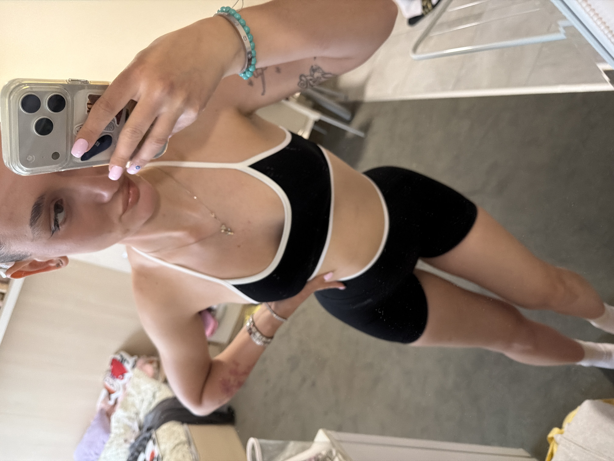 A woman is taking a selfie in front of a mirror. She is wearing a black and white sports bra and black shorts. The background includes a bed with bedding, a metal clothes rack, and a tiled floor. The location cannot be determined to be Sofia, Bulgaria from the information provided.