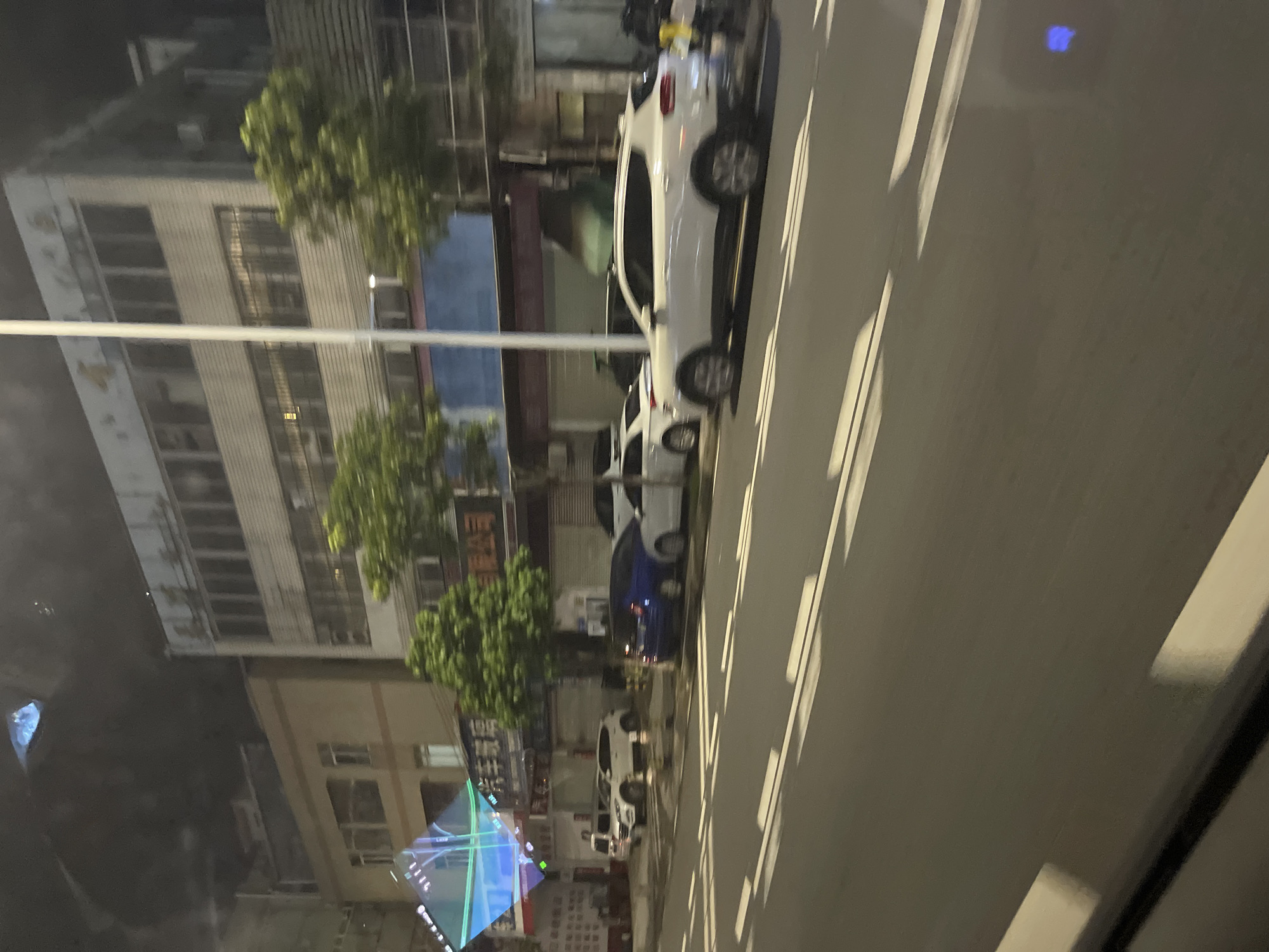 The image shows a street scene at night in Panyu District, China. In the foreground, a dark asphalt road with white lane markings is visible. The markings appear to be slightly blurred, suggesting motion. On the right side of the road, several cars are parked. A white SUV is prominently featured, followed by a blue sedan and another white car. Behind the parked cars, a building with multiple stories is visible. The building has a light-colored facade and appears to be aged, with some visible wear and tear. Windows are present on all levels. Trees with green foliage are planted along the sidewalk in front of the building. In the background, the sky is dark, indicating nighttime. A tall white pole, likely a street light or utility pole, stands vertically in the middle of the scene. To the far left, a triangular, illuminated structure with blue and green lights is partially visible, suggesting it might be a sign or decorative element. The overall impression is of a commercial or residential street in a Chinese city at night.
