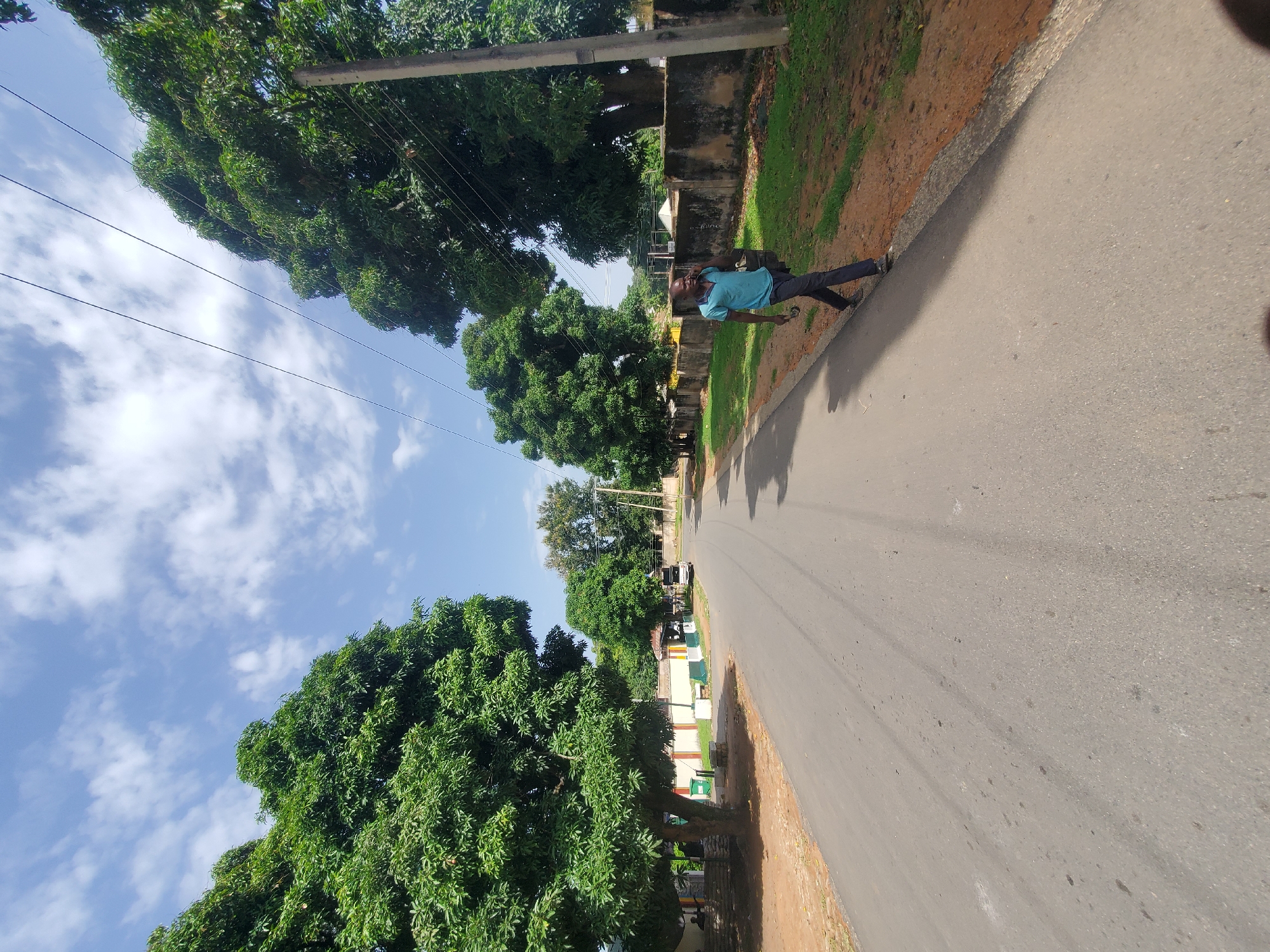 A person is walking down a paved road in Jos, Nigeria, on a bright, cloudy day. The road is lined with trees and a low wall. The person is wearing a blue shirt and dark pants, carrying a bag. A white vehicle is parked on the side of the road in the distance. The sky is blue with scattered white clouds. The environment appears to be a residential or semi-rural area within the city.