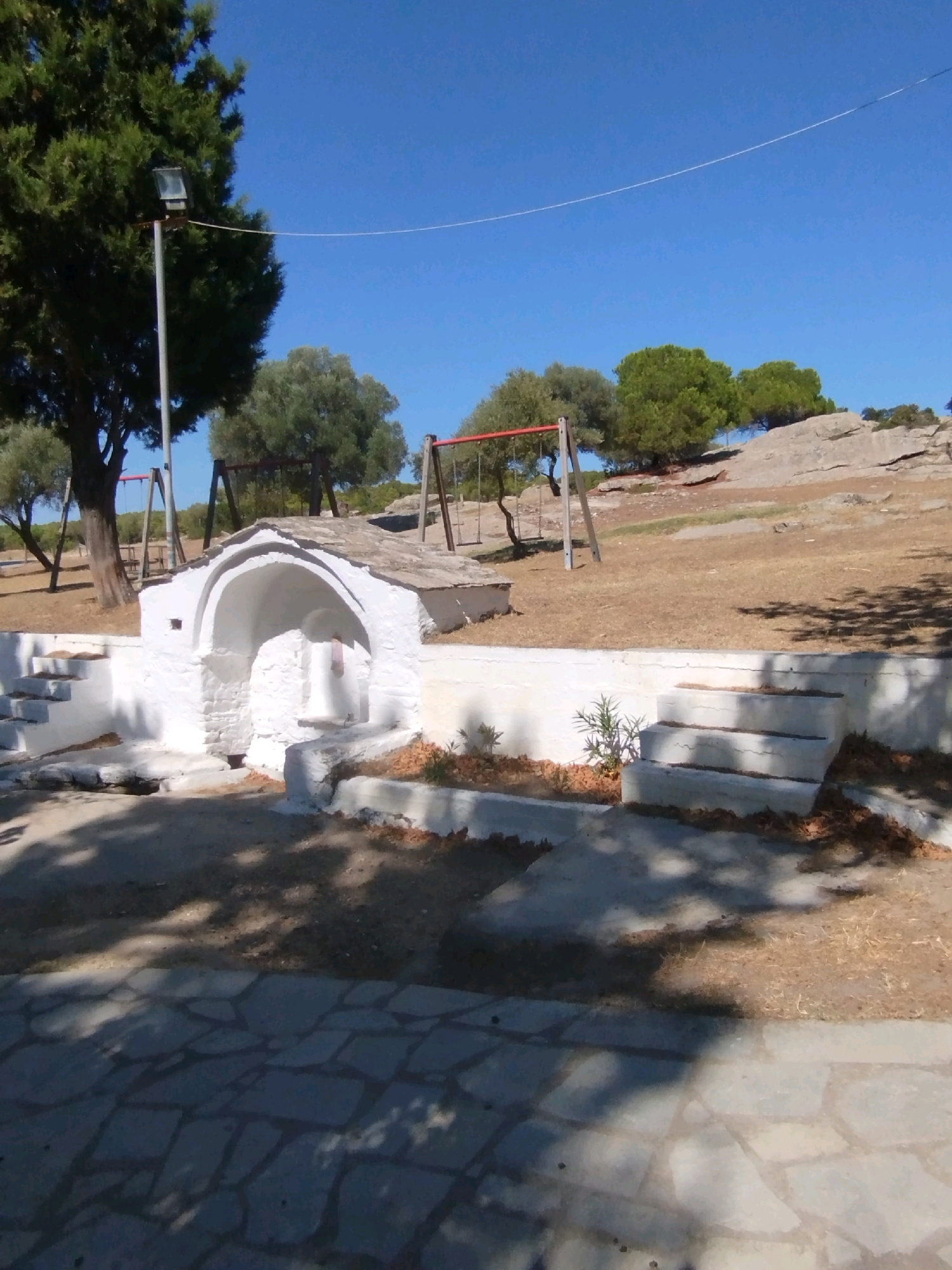 The image depicts an outdoor scene on a clear, sunny day. In the foreground, a paved area with irregularly shaped stones is visible, with shadows of trees cast upon it. To the right, a set of white concrete stairs leads upwards. Behind the stairs and to the left of them, a white structure with an arched opening and a stone roof stands. This structure appears to be a fountain or a small shrine. To the left of the structure, more white stairs ascend.
In the middle ground, dry, brown grass covers the ground. Several swings with red frames are set up, suggesting a playground area. A tall tree with dark green foliage stands to the left, with a floodlight attached to a pole near its base. Other trees with lighter green foliage are scattered further back. The background consists of a rocky, brown hillside under a clear blue sky.
The image is a photograph taken from a slightly elevated, static perspective. There are no people or animals visible in the frame. The scene suggests a quiet, possibly rural or park-like setting in the Sithonia Municipal Unit, Greece.
