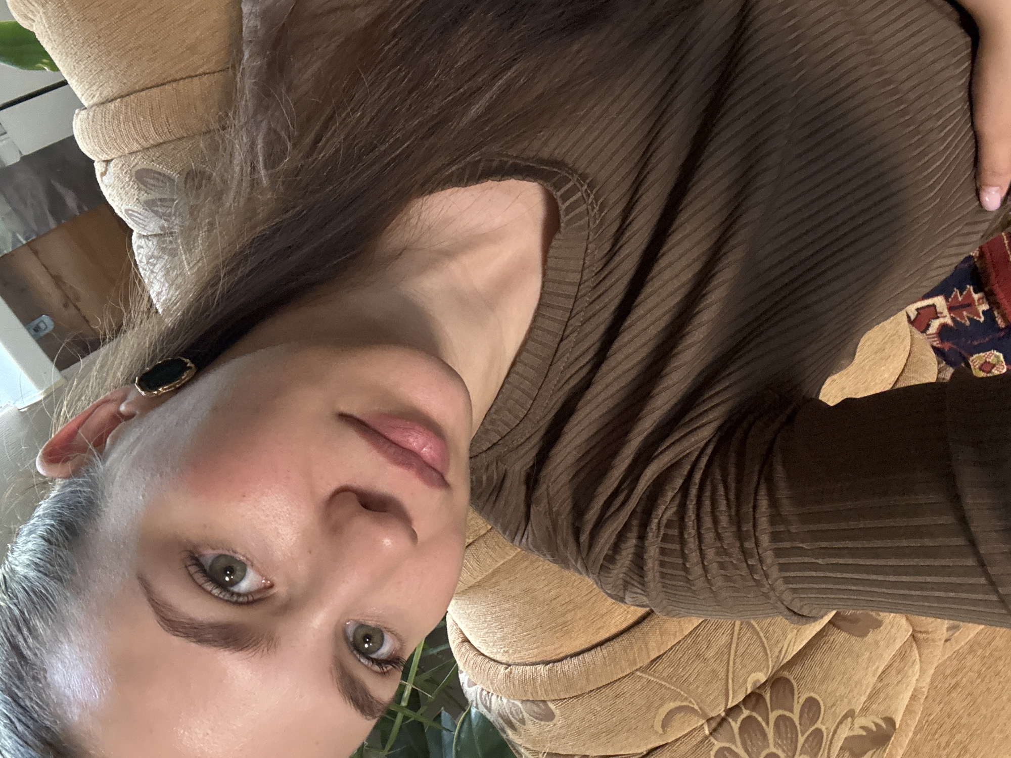 A close-up portrait, oriented diagonally, features a person, likely a woman, situated indoors in Nizhnyaya Gar', Russia. The subject looks directly at the camera with a neutral to slightly contemplative expression. She has green eyes, naturally pink lips, and long, dark brown hair, with visible gray strands around her temples and part.
She is dressed in a dark brown, ribbed long-sleeved top. A dark, possibly black or dark green, earring with a gold-colored rim is visible on her left ear. A manicured hand with light pink nail polish is partially visible on her right side.
The background consists primarily of a light brown or beige upholstered couch or chair featuring a patterned fabric with floral or leaf-like designs. A fragment of a geometrically patterned rug or textile, displaying colors like red, blue, white, and brown, is visible to the right. Green foliage from an indoor plant can be seen in the top and bottom left sections of the frame. The lighting suggests it is daytime indoors. No text is visible in the image.