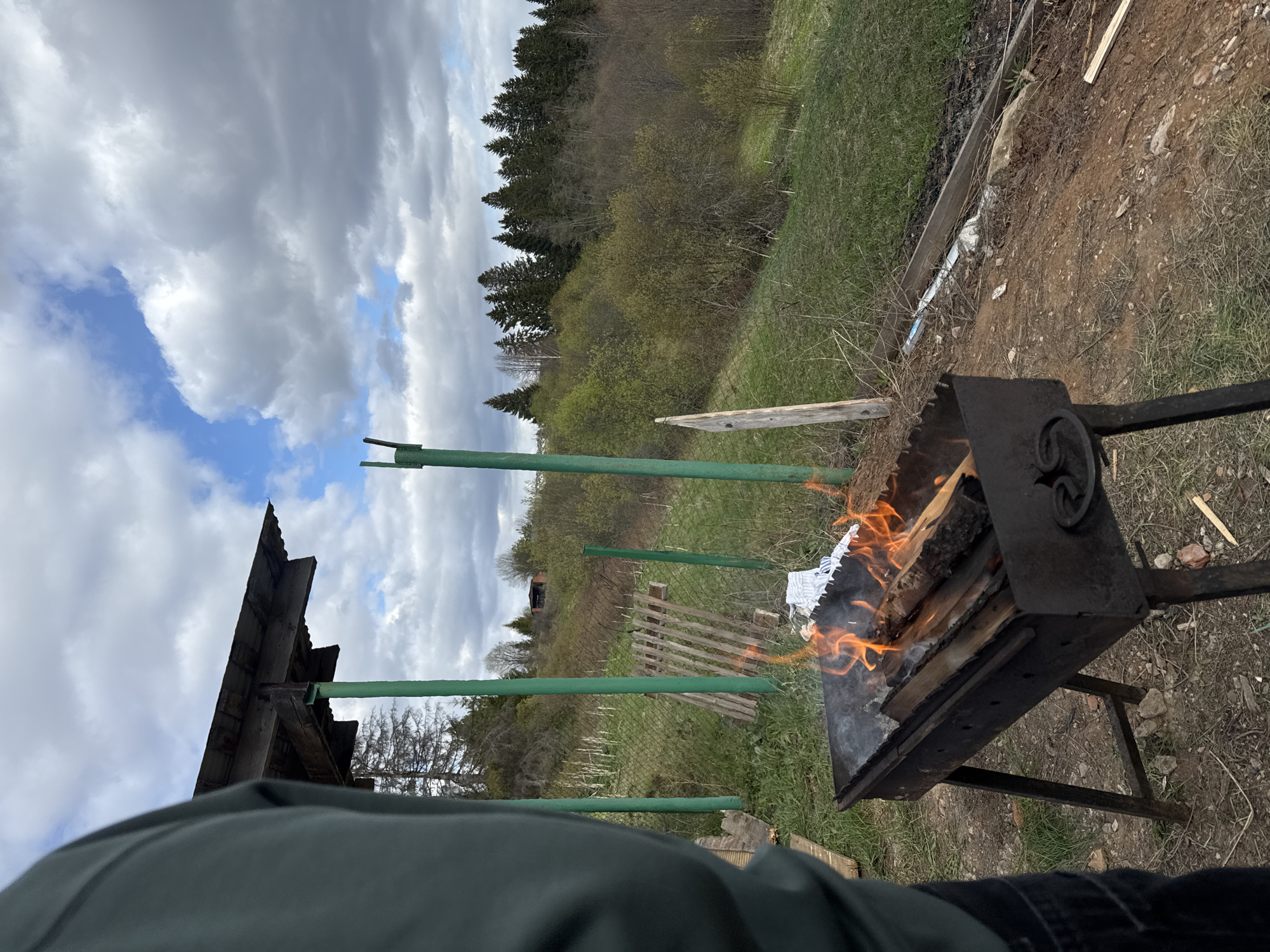 The image depicts a rural scene, potentially in Abrosy, Russia. A metal barbecue grill is positioned on grassy terrain, with a fire burning inside. To the left, a partially visible structure, possibly a shed, stands with green painted supports. Behind the grill, there's a wire fence and a dilapidated wooden gate leading into a field or meadow with distant trees. The sky overhead is cloudy with patches of blue visible. A person's torso, clad in a green jacket and black pants, is partially visible at the bottom of the frame, suggesting the image was taken from a first-person perspective.