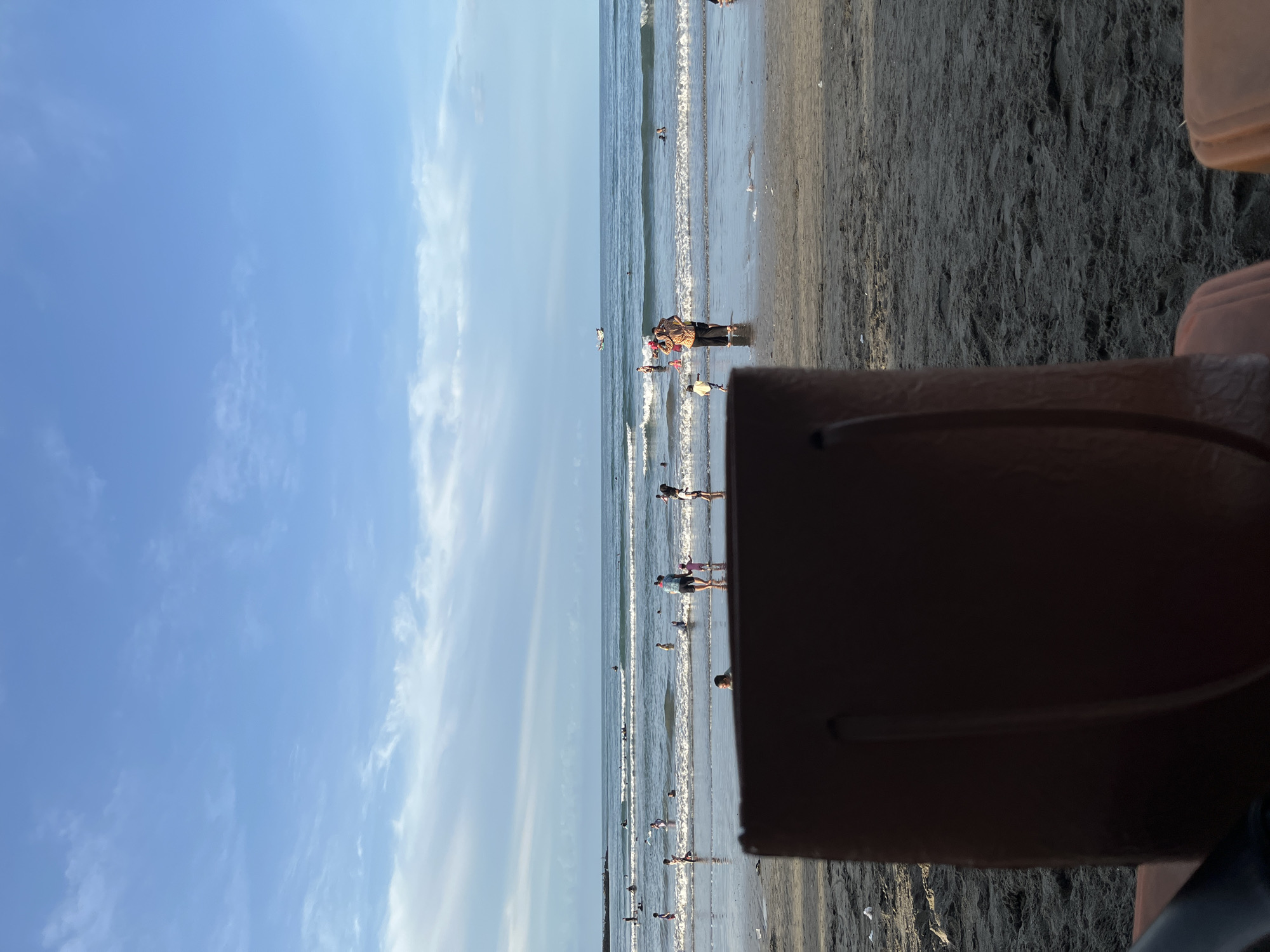 The image shows a beach scene in Lhokseumawe, Indonesia. The perspective is from a seated position, with a chair partially obscuring the view. The beach has dark sand, and people are visible along the shoreline and in the water. The sea has small waves. The sky is blue with scattered clouds.