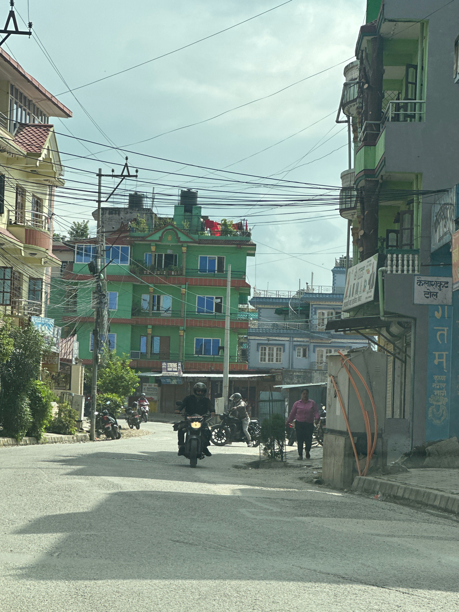 The image depicts a street scene in Kageshwori Manohara, Nepal. A multi-story green building with red accents and multiple blue-windowed floors dominates the center of the frame, with a similar yellow building on the left. A gray building with balconies, some featuring intricate railings, stands to the right, with a large tree trunk growing through one of its balconies. Overhead, a complex network of electrical wires crisscrosses the sky. On the street, several people are visible. In the foreground, a person rides a motorcycle, facing away from the camera. Further down the road, another individual on a motorcycle is seen, along with a person in a pink shirt and dark pants walking. Parked motorcycles are present along the left side of the street. The road surface is paved and shows signs of wear, with shadows cast across it from the buildings. The sky is overcast. Signs with text in a script that appears to be Nepali are affixed to some of the buildings.