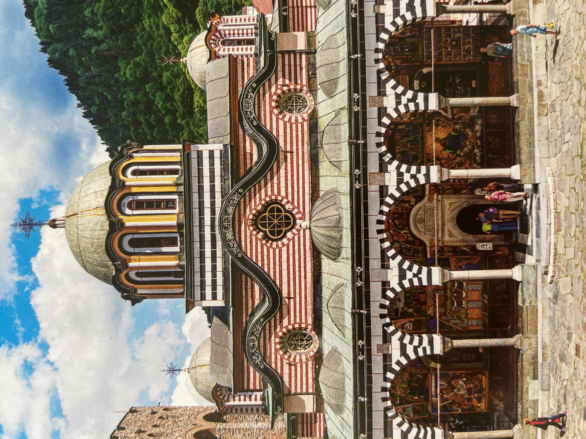 The image displays the Rila Monastery in Bulgaria, an Eastern Orthodox monastery renowned for its distinctive and colorful architecture. The perspective is from the ground, looking up at the elaborate main church building and its surrounding structures.
The central edifice features multiple domes, including a large, prominent one with a greyish-green hue, each topped with an ornate metallic cross. The building's facades are richly decorated: one section showcases vertical yellow, orange, and brown stripes around arched windows, while another section is adorned with striking vertical red and white stripes, accentuated by a wavy black decorative band and circular windows with intricate geometric grilles.
To the right, a multi-arched portico or cloister is visible, featuring a bold pattern of black and white stripes on its arches, supported by robust white columns. Within these arched sections, vibrant and detailed frescoes depicting religious narratives are painted on the walls.
The monastery is set against a dramatic backdrop of a dense, green forested mountain range that rises steeply behind the complex, contrasting with a bright blue sky dotted with scattered white clouds. In the foreground, a spacious courtyard paved with irregular grey cobblestones extends in front of the monastery.
Several people, dressed in casual attire, are scattered across the cobblestone courtyard. They appear to be visitors or tourists, observing the monastery's architecture, walking, or standing. The scene depicts a lively but reverent atmosphere of exploration at a significant religious and cultural site.
The image captures the scene during daytime, under clear weather conditions, indicated by the bright and even lighting. There is no visible text in the image.