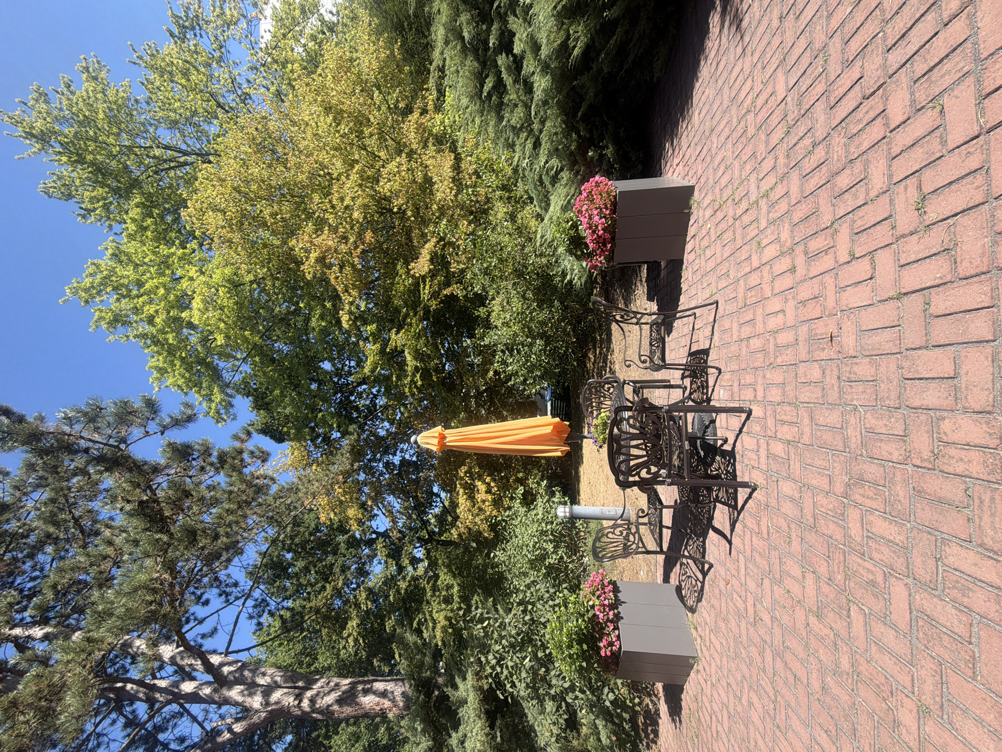An outdoor patio area paved with red brick is furnished with a metal table and four chairs. An orange umbrella stands open behind the table, casting a shadow. Two gray planters filled with pink flowers are positioned on either side of the seating area. Behind the patio, a variety of trees and shrubs form a lush green backdrop. The scene is illuminated by bright sunlight, and the sky is clear blue. There are no people visible in the image. The location is the city of Dusseldorf, Germany.