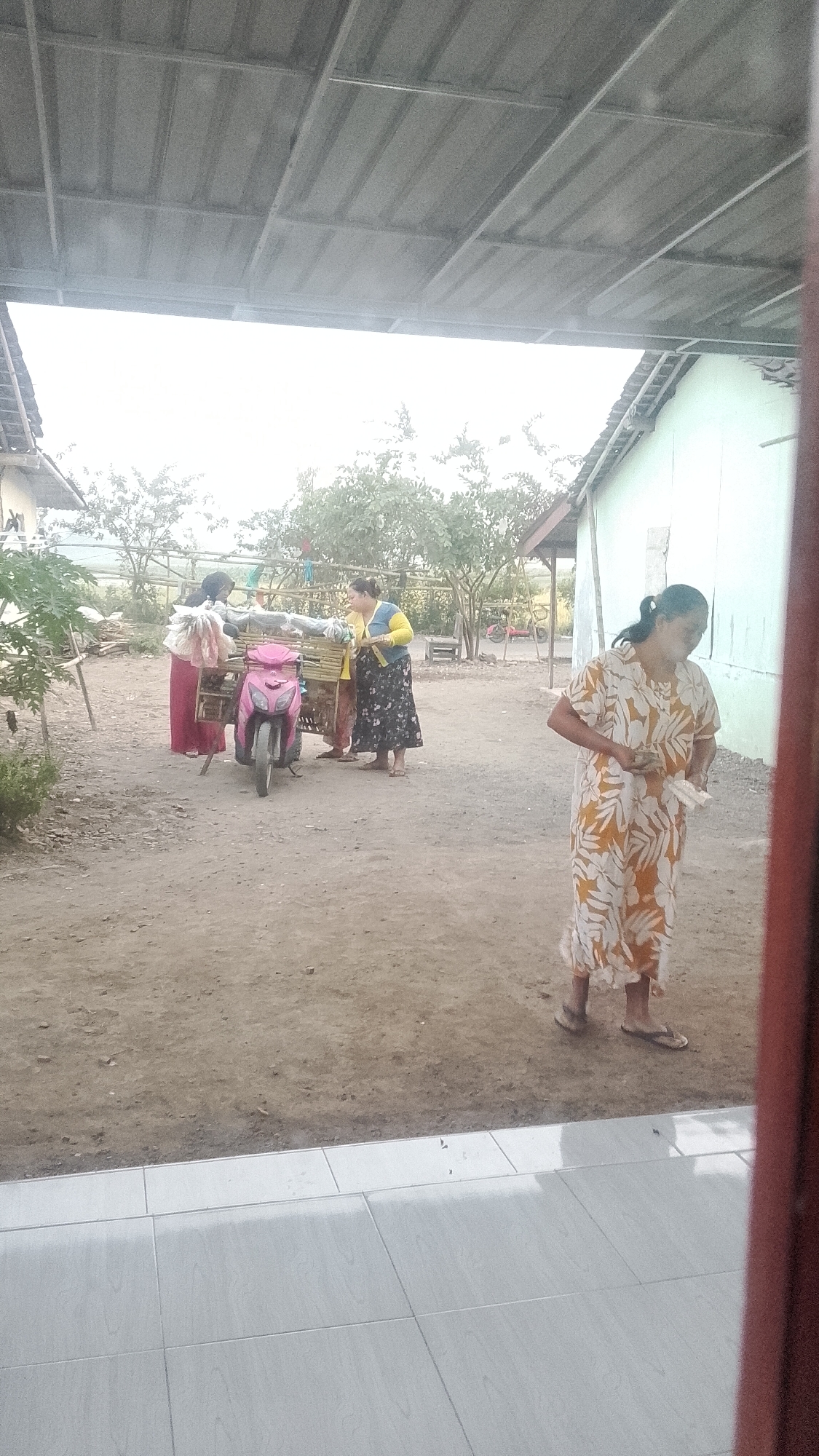 An outdoor scene in Karangkedawung, Indonesia shows three individuals interacting near a pink scooter with a wooden cart attached. One woman, wearing a patterned yellow and white dress and flip-flops, stands to the right holding several white items. To the left, two other women, one in a red dress and the other in a yellow and blue top with a floral skirt, stand near the scooter and cart, which appears to be laden with goods. The ground is dusty dirt, and a tiled patio is visible in the foreground. In the background, a building with a light-colored wall and a corrugated metal roof can be seen, along with trees and vegetation. The overall impression is of a rural or semi-rural setting.
