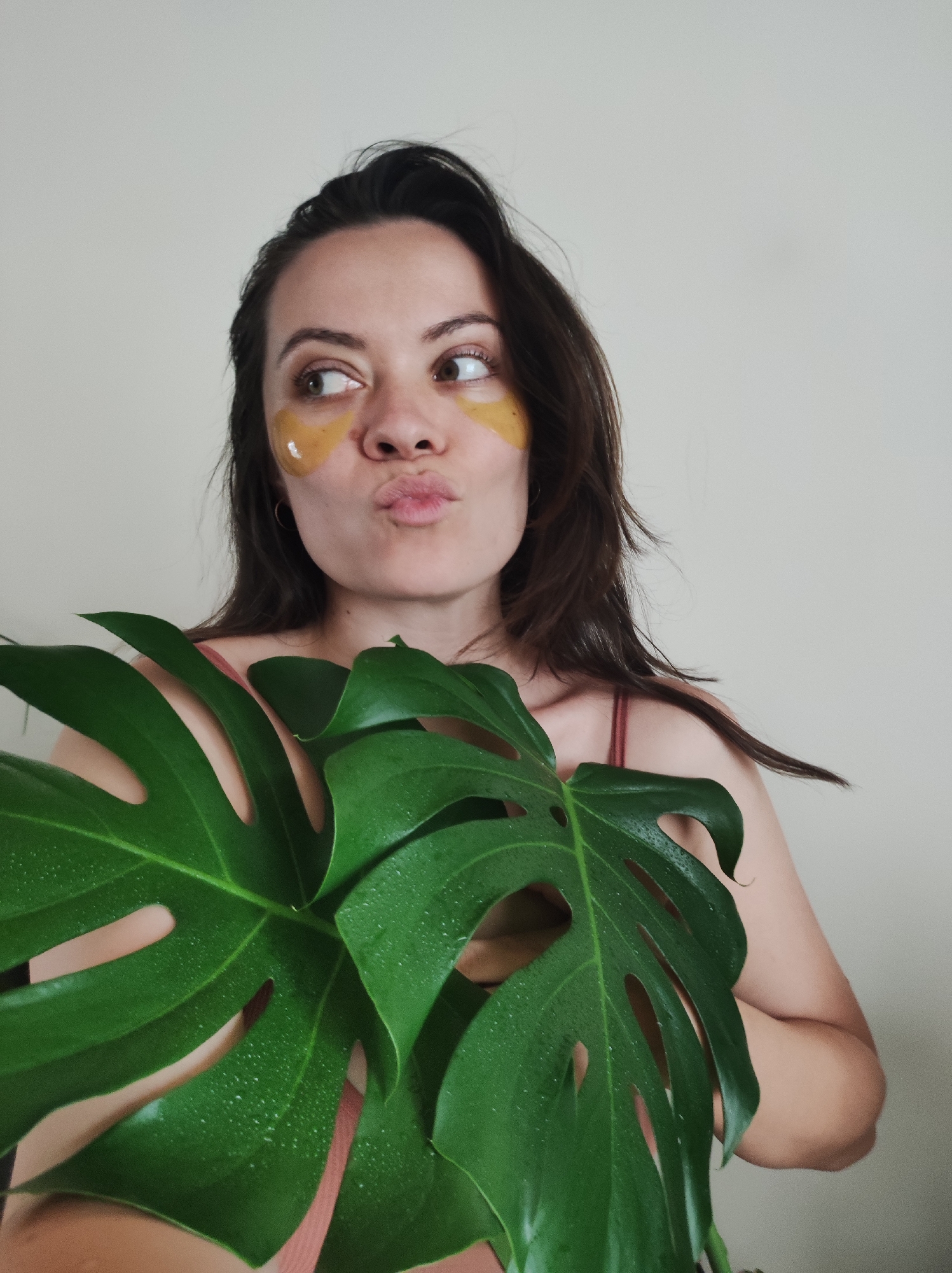 A woman poses with a Monstera deliciosa leaf in front of a plain white wall. She has eye patches on and is pouting her lips. Her hair is dark brown. The plant leaf has water droplets on it. The woman is wearing a pink top. The location is Unknown, Unknown.