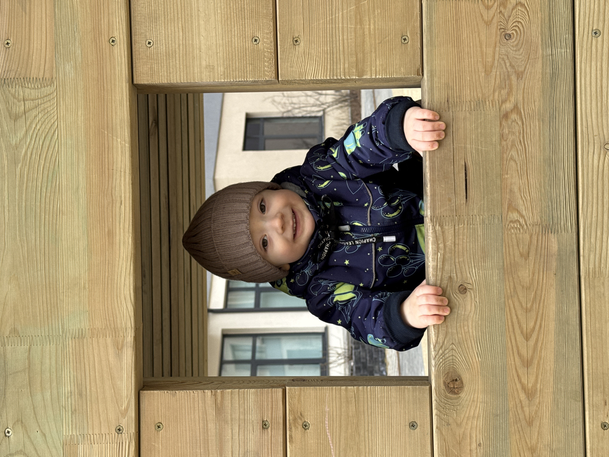 A young child wearing a brown knit hat and a dark blue jacket with space-themed patterns is smiling and looking directly at the camera. The child is positioned within a square opening in a wooden structure, possibly a playhouse or climbing frame, constructed from light-colored wood planks. The child's hands are visible, gripping the edges of the wooden opening.
Through the opening, a blurred background reveals a multi-story building with dark-framed windows and a bare tree.
The location is in the city of Izhevsk, Russia.