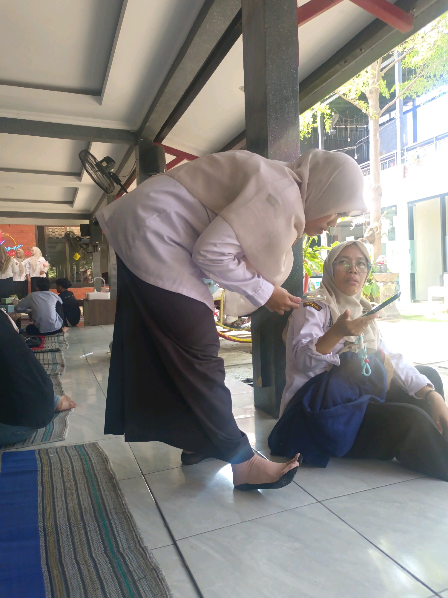 In Kerangkulon, Indonesia, two women are interacting outdoors on a tiled floor. The woman on the left is wearing a light-colored hijab and a long, dark skirt, with her left foot visible. The woman on the right is also wearing a light-colored hijab and a white shirt with badges, and appears to be looking at a mobile phone she holds in her right hand. She is seated with a dark blue cloth on her lap. In the background, another group of people are seated on woven mats. A ceiling fan is visible mounted on a pillar to the left. Trees and a building are seen in the background.