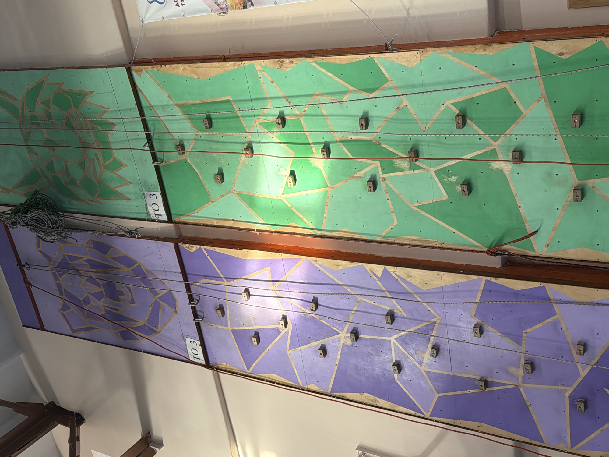 The image depicts a climbing wall inside a building, likely a gymnasium or recreational facility. The wall is divided into two distinct sections: the upper section is painted in shades of green with geometric patterns, and the lower section is purple with similar patterns. Both sections have evenly spaced climbing holds. There is rope on the upper left of the green climbing wall. The location in the city of Berezniki, Russia cannot be determined from the image.