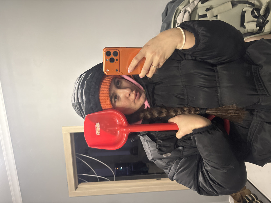 A woman in a black puffer coat, orange beanie, and pink hoodie takes a selfie in a mirror. She holds a red plastic shovel across her chest. The phone in her hand has an orange case. She is looking at the camera. Her hair is in a braid. A window is visible in the background, showing a dark night sky.