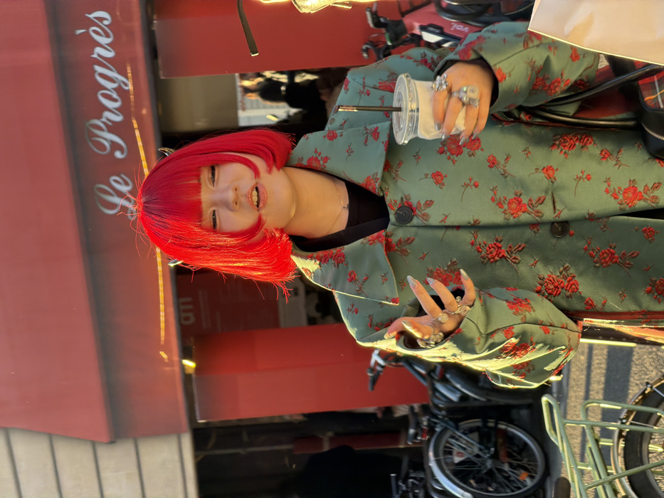 A person with bright red hair wears a green floral patterned jacket, holding a drink with a straw. They stand outside a building with "le Progrès" written on it. The person has long nails and wears multiple rings on their fingers. Bicycles are visible in the background, suggesting a street scene. The picture is rotated 90 degrees clockwise.