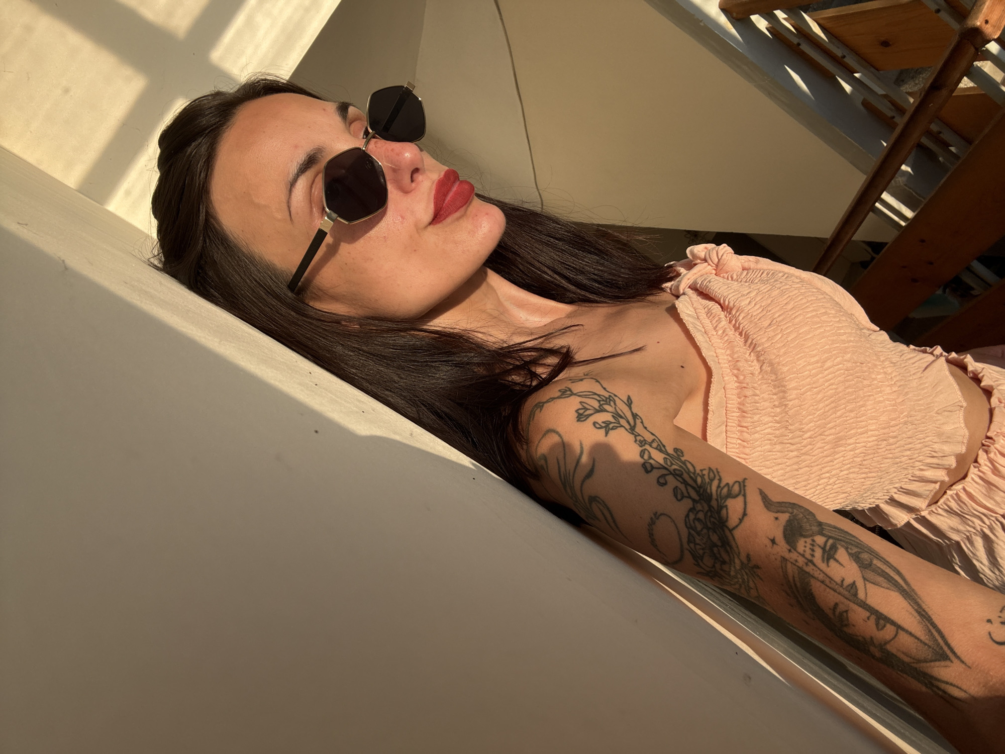 A young woman with dark hair, wearing a pink smocked top and hexagonal sunglasses, is leaning against a light-colored wall. Sunlight streams in from the side, casting shadows on the wall and the woman's face and arm. She has tattoos on her left arm, including a depiction of two faces and floral elements. The background shows a wooden staircase with some sunlight filtering through. The scene appears to be an interior staircase or hallway in Pleven, Bulgaria.
