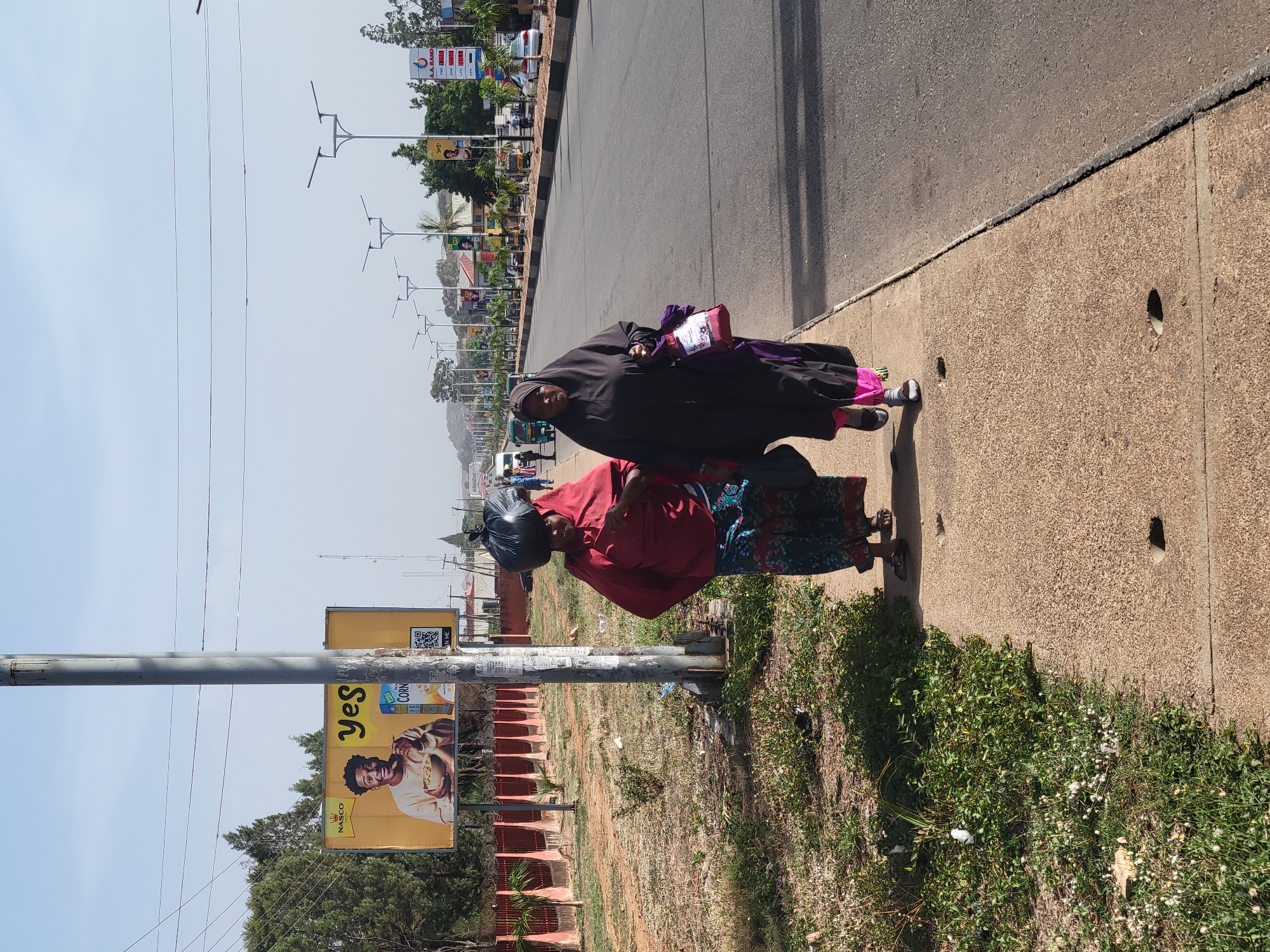 Two women walk on a sidewalk next to a road in Jos, Nigeria. One woman wears a black hijab and carries a purple bag. The other woman wears a red top and a patterned skirt, with a black head covering. A utility pole stands beside them, featuring an advertisement for "Yes Corn Flakes." The road is visible, with cars and buildings in the distance. Grass and vegetation grow along the edge of the sidewalk. The sky is pale and clear.