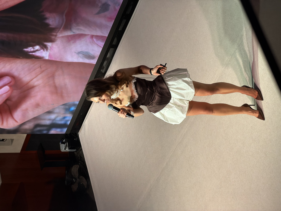 A female individual stands on a light-colored stage in an indoor event space in Moscow, Russia. She is speaking into a dark-colored microphone held in her left hand and holds a small black remote connected to a beaded strap in her right hand. She is dressed in a strapless dark brown corset with white lace trim, a voluminous short white skirt, and brown high-heeled pointed-toe shoes. Multiple pearl necklaces, including a choker, and a pearl bracelet accessorize her attire. Behind her, a large LED screen displays a magnified image of a human hand with pink manicured fingernails and a portion of a person's torso. To the left side of the stage, a dark high-top table, a dark floor fan, and a piece of electronic equipment are visible against a dark wall.