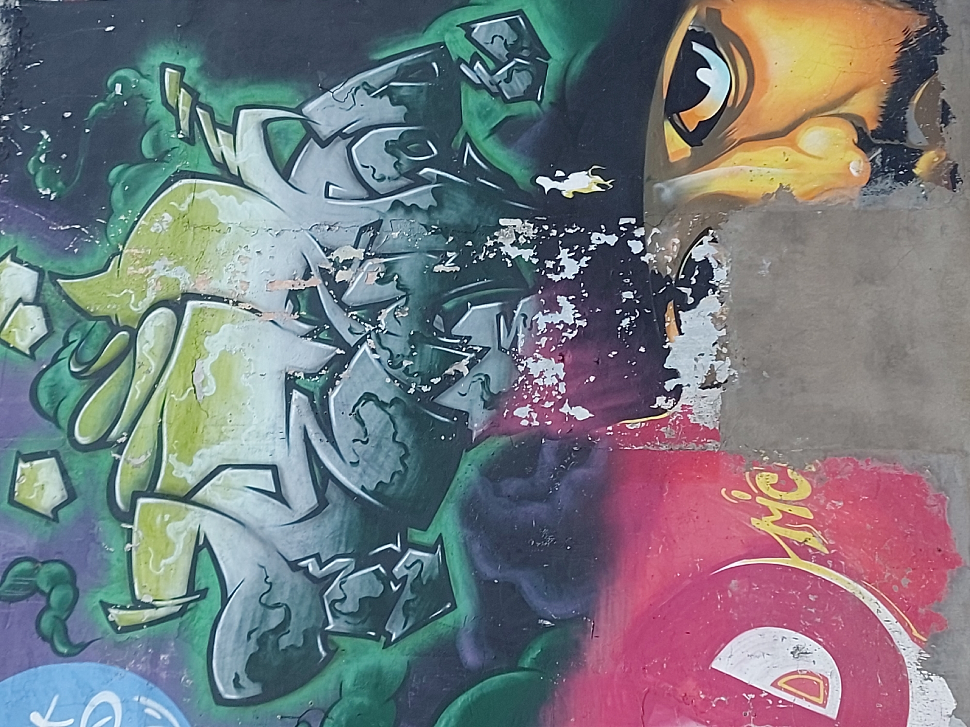 A close-up view of a decaying graffiti mural on a concrete wall. The mural exhibits multiple layers and styles. On the left, there's abstract green and grey graffiti with defined edges. Adjacent to it, there's a large cartoon eye, predominantly yellow and black. In the lower right, there's a red circle with a white segment and yellow lettering. The paint is peeling and flaking in multiple areas, revealing the underlying concrete. There are no people visible in the scene. This image was taken in Izhevsk, Russia.