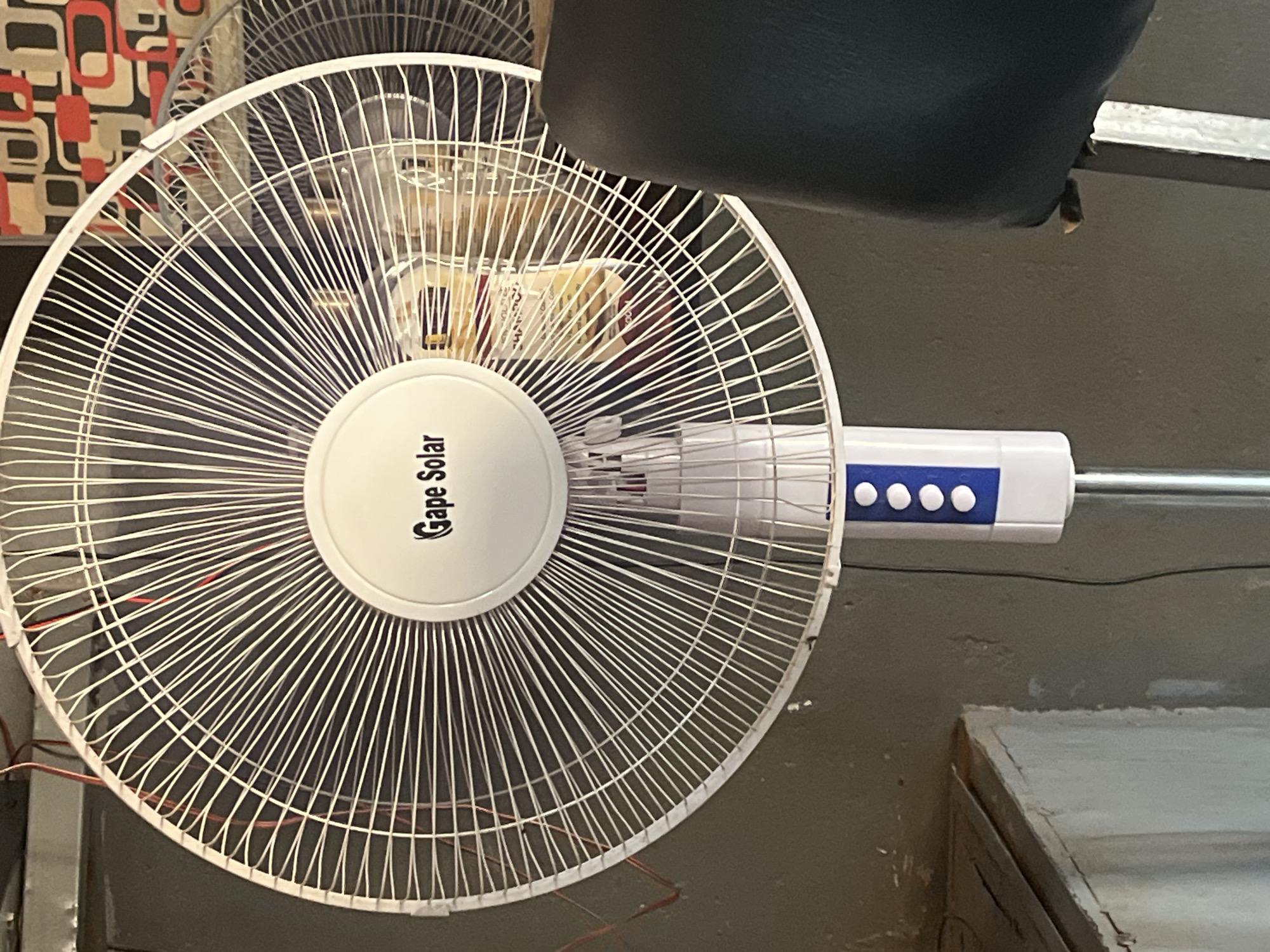 The image depicts a white "Gape Solar" brand electric fan with a metal cage and a white and blue control panel. The fan is in focus against a blurred background consisting of a wall and what appears to be a checkered object behind the fan. The location is stated to be Rigasa, Nigeria, though this information is not visually verifiable. No people are visible.
