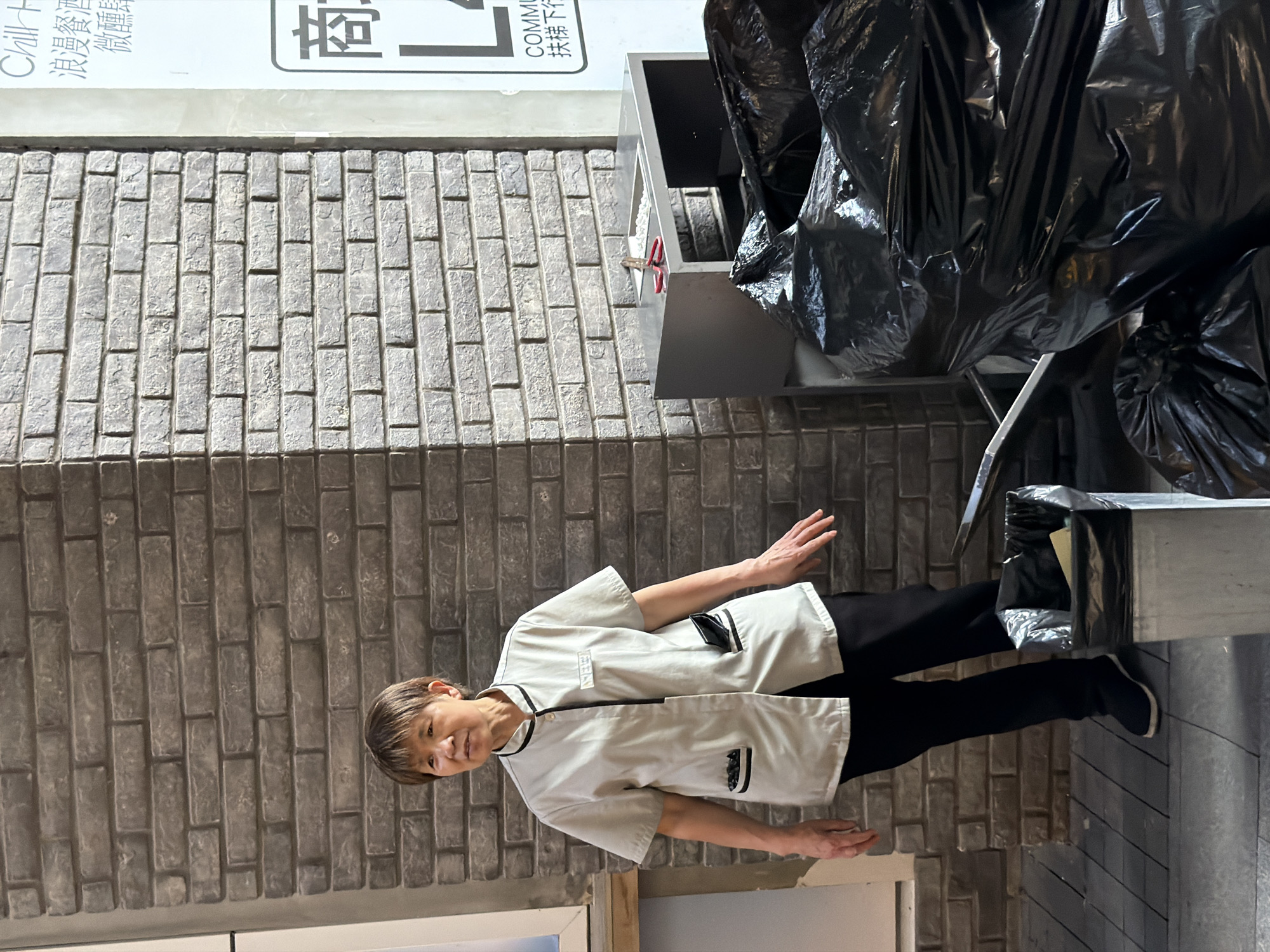 An older East Asian woman with short, dark hair stands in front of a brick wall and a trash receptacle. She is wearing a light-colored, short-sleeved uniform shirt with black trim and black pants. Her left hand is resting on her hip, and her right arm is slightly extended. To her right, several large black trash bags are piled high, and a metal trash can with a black liner is visible. A small silver desk with red scissors on top is positioned between the brick wall and the trash bags. A sign with Chinese characters is affixed to the wall in the upper right quadrant of the image. The environment appears to be an outdoor urban setting, possibly a back alley or service area, in Nan'an District, China.