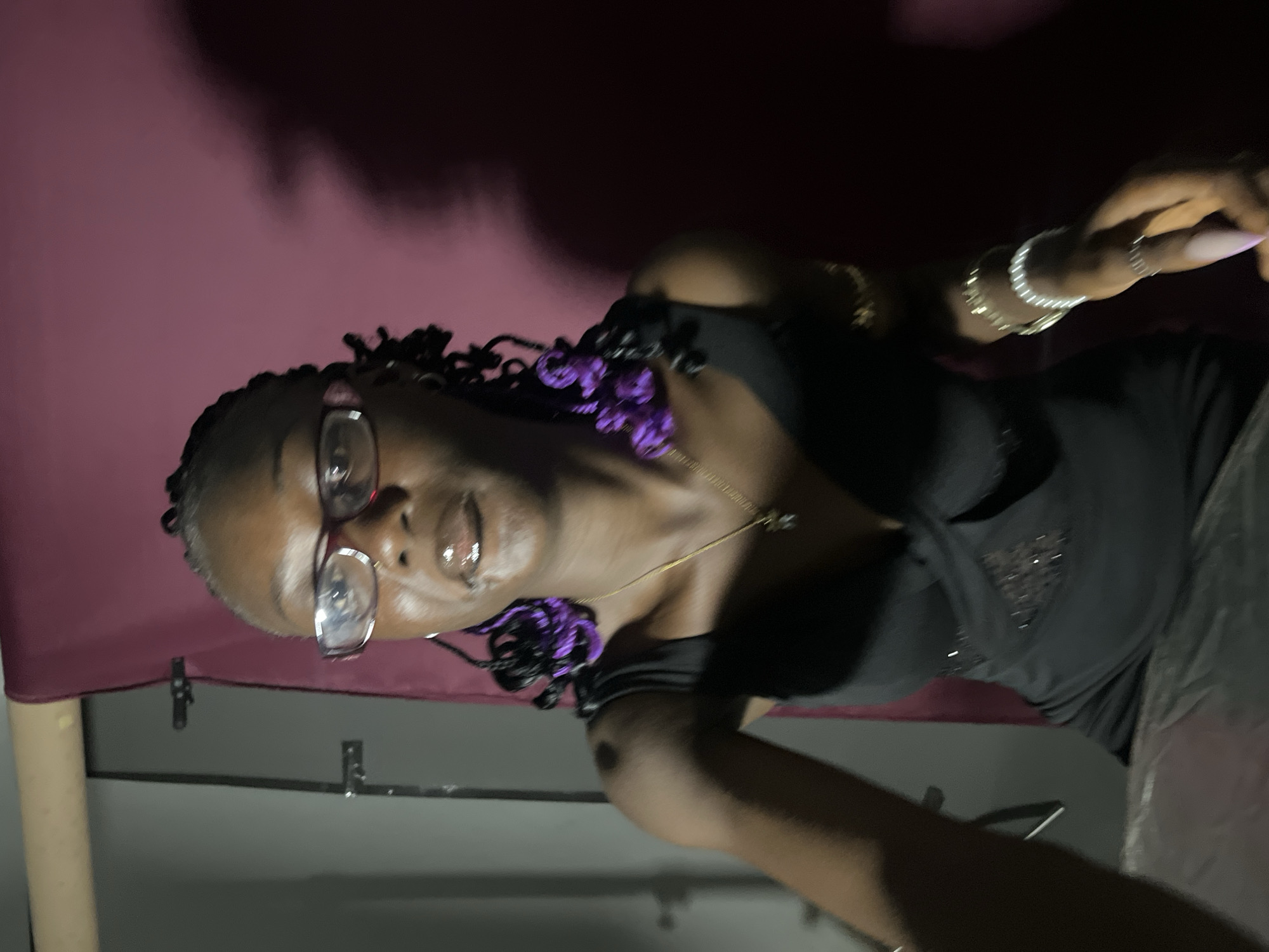 A medium shot captures a Black woman with braided hair and purple extensions from the chest up, facing forward and looking directly at the camera. She is wearing glasses with pink rims, a black dress with a V-neckline and decorative accents, a gold necklace, and several bracelets on her wrists. Her lips are glossy and appear slightly parted. The background consists of a maroon fabric backdrop with a dark shadow cast on the right side, and a beige-colored cylindrical object and gray wall on the left. The setting suggests an indoor studio or photo booth environment, potentially in Gura Top, Nigeria, given the prompt's context.