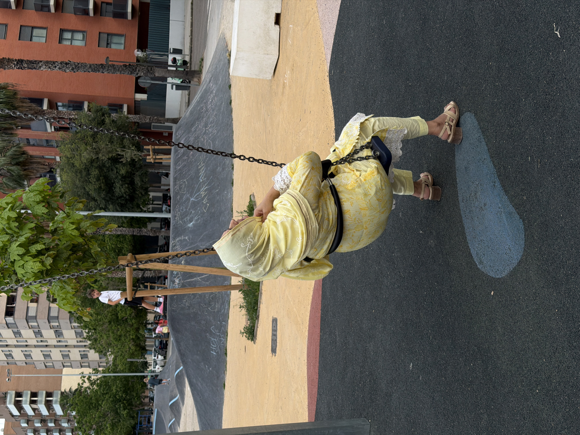 A person wearing a yellow outfit and beige sandals is swinging on a swing set in an outdoor playground. The playground surface is a mixture of black and tan surfaces. Behind the swing set, there is a cityscape that includes apartment buildings and trees. The photograph was taken in Valencia, Spain.