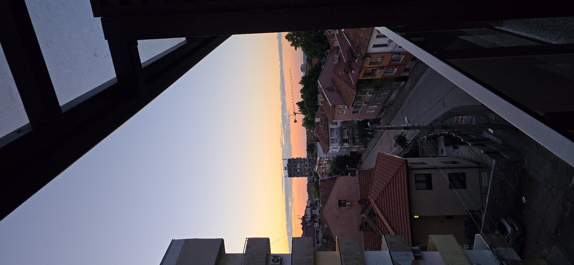 The image captures a dusk scene overlooking a residential street in a city. In the foreground, a dark balcony railing and overhang frame the view. Beyond, a cluster of buildings with orange-tiled roofs line a winding street. A multi-story apartment building is visible in the distance, and a construction crane stands out against the twilight sky, which transitions from a pale blue at the top to a warm orange and yellow band near the horizon, dotted with faint clouds. The overall impression is of a quiet urban evening.