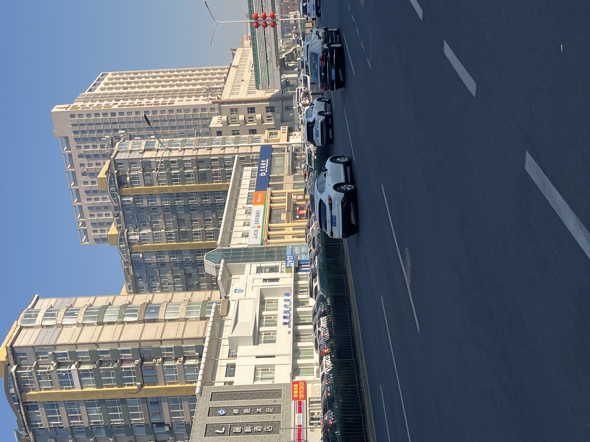 The image depicts a street scene in Changchun City, China, featuring several buildings, traffic, and street signage. The buildings are mostly high-rise structures with a modern architectural style, including residential and commercial properties. Along the street, there are vehicles parked or driving, including cars and potentially other types of automobiles. The street also features lane markings. Signage is visible for businesses and services, including financial institutions and retail establishments. Overall, the image captures a typical urban environment in Changchun City, China.