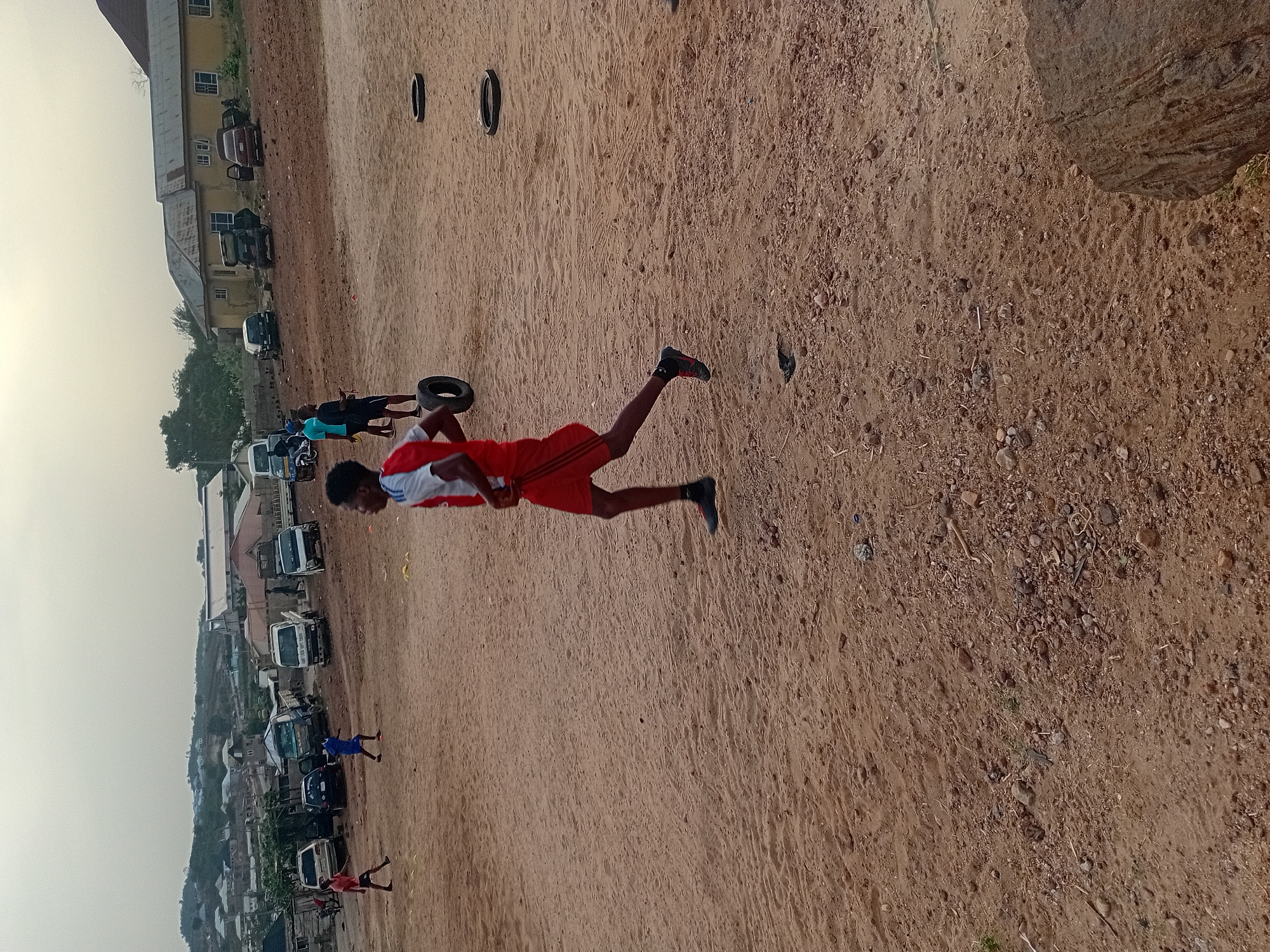 The image depicts an outdoor scene, likely a training or exercise ground, under an overcast sky. The primary subject is a dark-skinned male in the foreground, captured mid-action. He is wearing a red and white athletic t-shirt, red shorts, and black athletic shoes. He is positioned in a jumping or running stance, with one leg extended forward and the other bent at the knee, appearing to be in mid-air. He holds a black car tire with both hands, suggesting he is either lifting, throwing, or jumping over it as part of a workout.
The setting is a large expanse of reddish-brown dirt ground, scattered with small stones and debris, indicating a dry, unpaved area. In the mid-ground, several other individuals are visible, also engaged in activity or observing. One person wears a blue top and black shorts, and another in dark attire is near another black tire. Multiple black tires are strewn across the dirt field, reinforcing the impression of a training session or obstacle course.
In the background, a row of low-rise buildings with light-colored walls and dark roofs can be seen, suggesting a residential or light commercial area adjacent to the training ground. Several vehicles, including vans and cars, are parked along the side of these buildings. Beyond the immediate structures, distant hills or further buildings are faintly visible under the hazy sky. The time of day appears to be daytime, possibly late afternoon, given the relatively soft and diffused lighting. There is no visible text in the image.