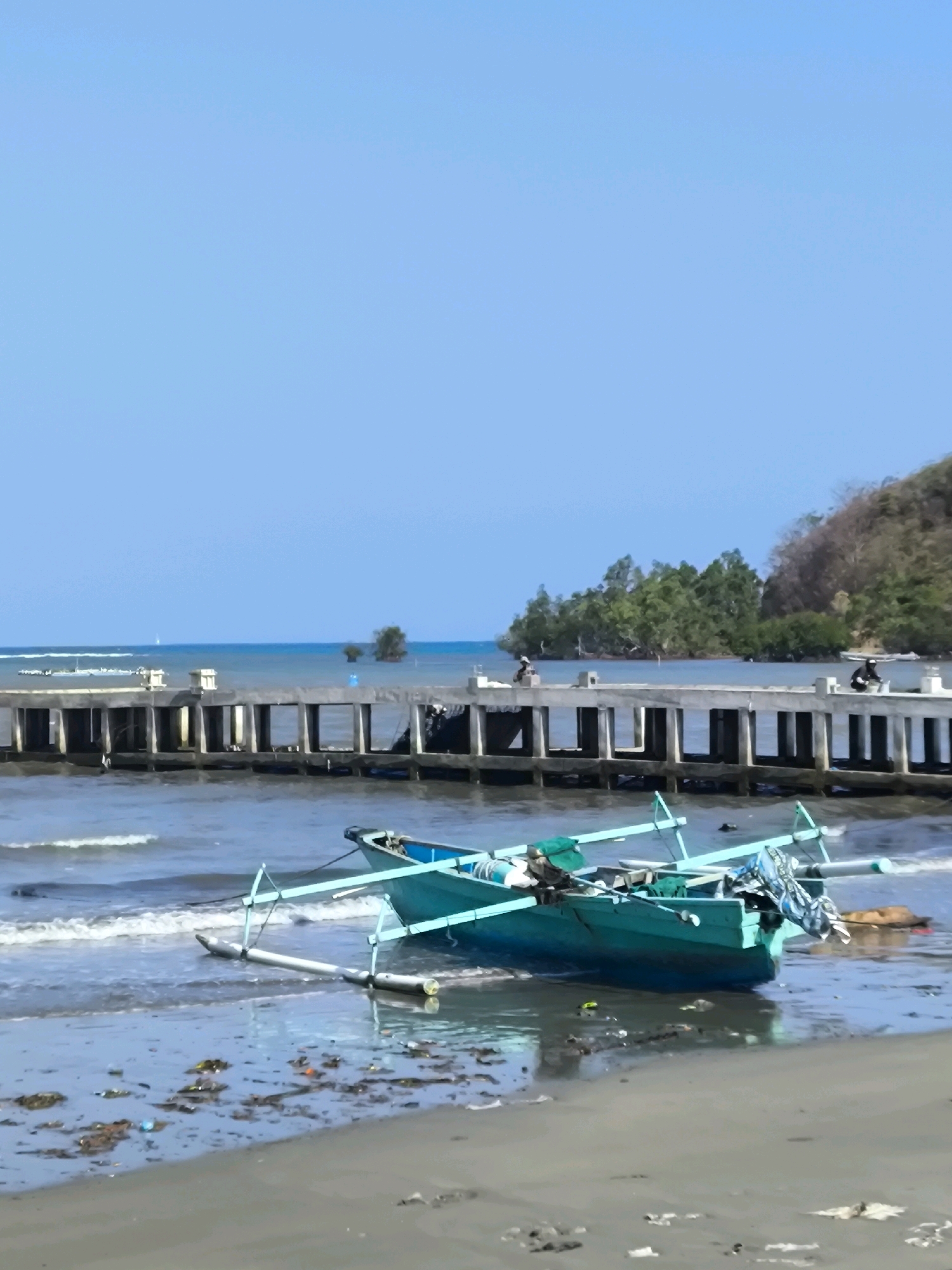 The image depicts a bright, sunny day at a beach with a clear blue sky and calm water. In the foreground, a turquoise-colored boat is partially submerged at the water's edge on a sandy shore littered with debris. The boat has outriggers and appears to be a fishing vessel. In the background, a long concrete pier extends into the ocean, with several people visible on it, some appearing to be engaged in activities. Beyond the pier, lush green trees and a hillside form the coastline. The scene suggests a tranquil coastal environment in Tilamuta, Indonesia, likely a fishing village or a popular beachside location.
