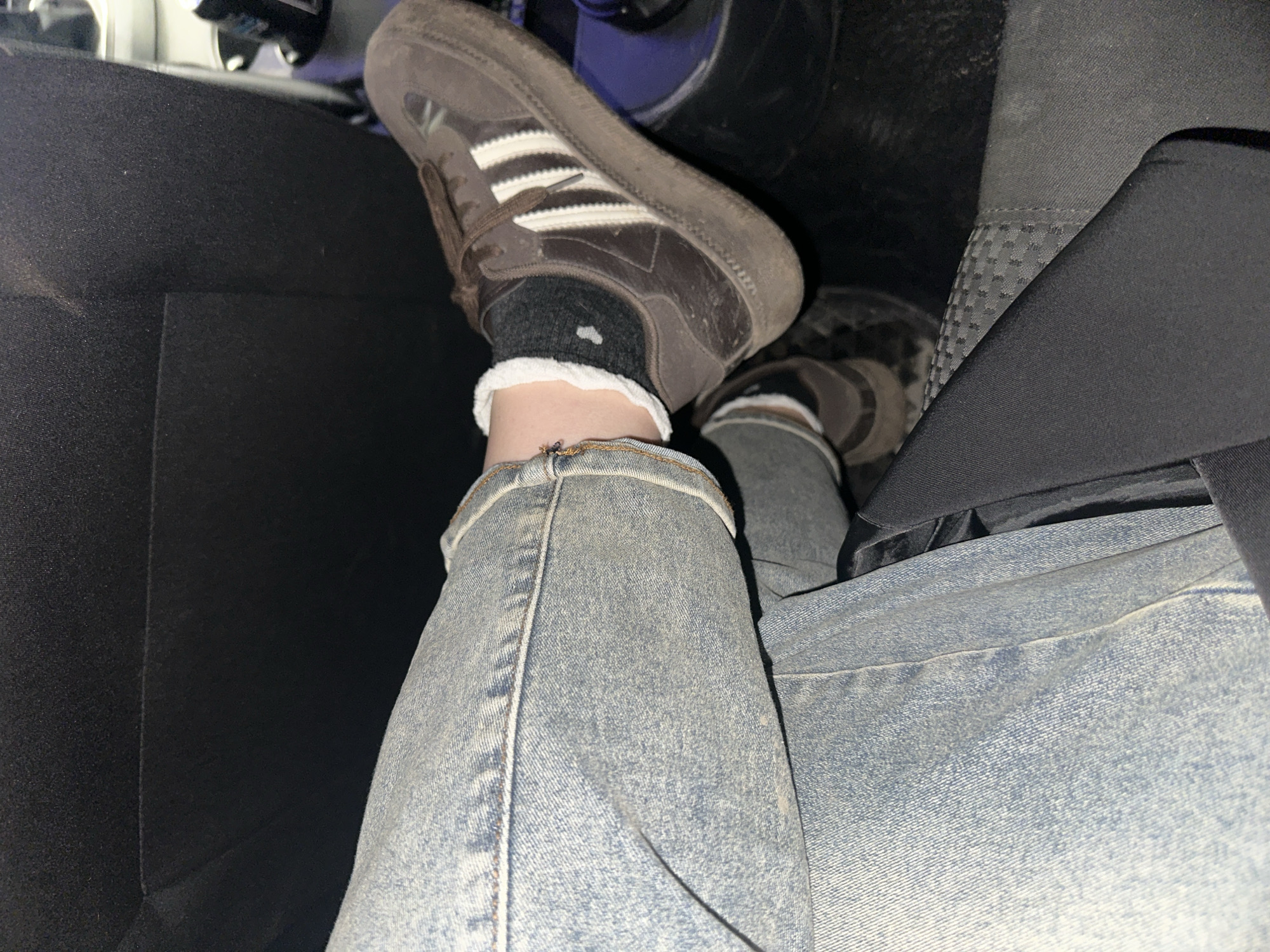 A close-up shot taken inside a vehicle shows a person's legs and feet. The person is wearing light-wash jeans, a dark grey sock with white trim, and a brown Adidas Samba shoe. The car's interior is visible, including the black upholstery of the seat and other parts of the car's interior. The background is dark and somewhat out of focus. The location is unknown.