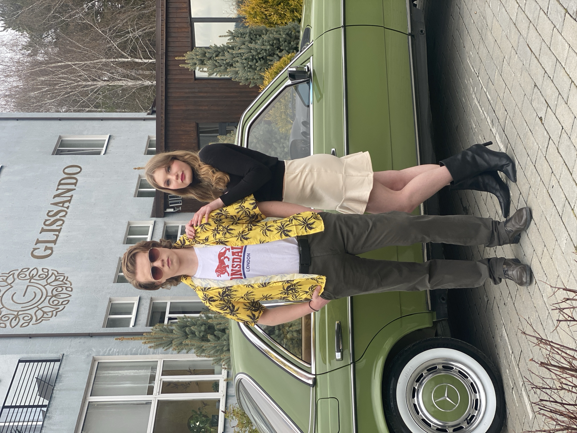 A young man and woman are posing next to a vintage green Mercedes-Benz car. The woman is fair-skinned with blonde hair, wearing a black turtleneck top and a beige miniskirt with black high-heeled boots. She is leaning against the car. The man has fair skin, light brown hair, and is wearing sunglasses. He is wearing a white t-shirt with the word "London" on it and a palm tree-print yellow Hawaiian shirt, with green pants and dark shoes. He is standing in front of the car. The car has a white-walled tire and a visible Mercedes-Benz logo on the hubcap.
The background includes a building with the word "GLISSANDO" vertically printed on the building. There are also evergreen bushes and trees. Based on the architectural style and signage, the location is in Yekaterinburg, Russia. The surface they stand on is paved with light-colored bricks.