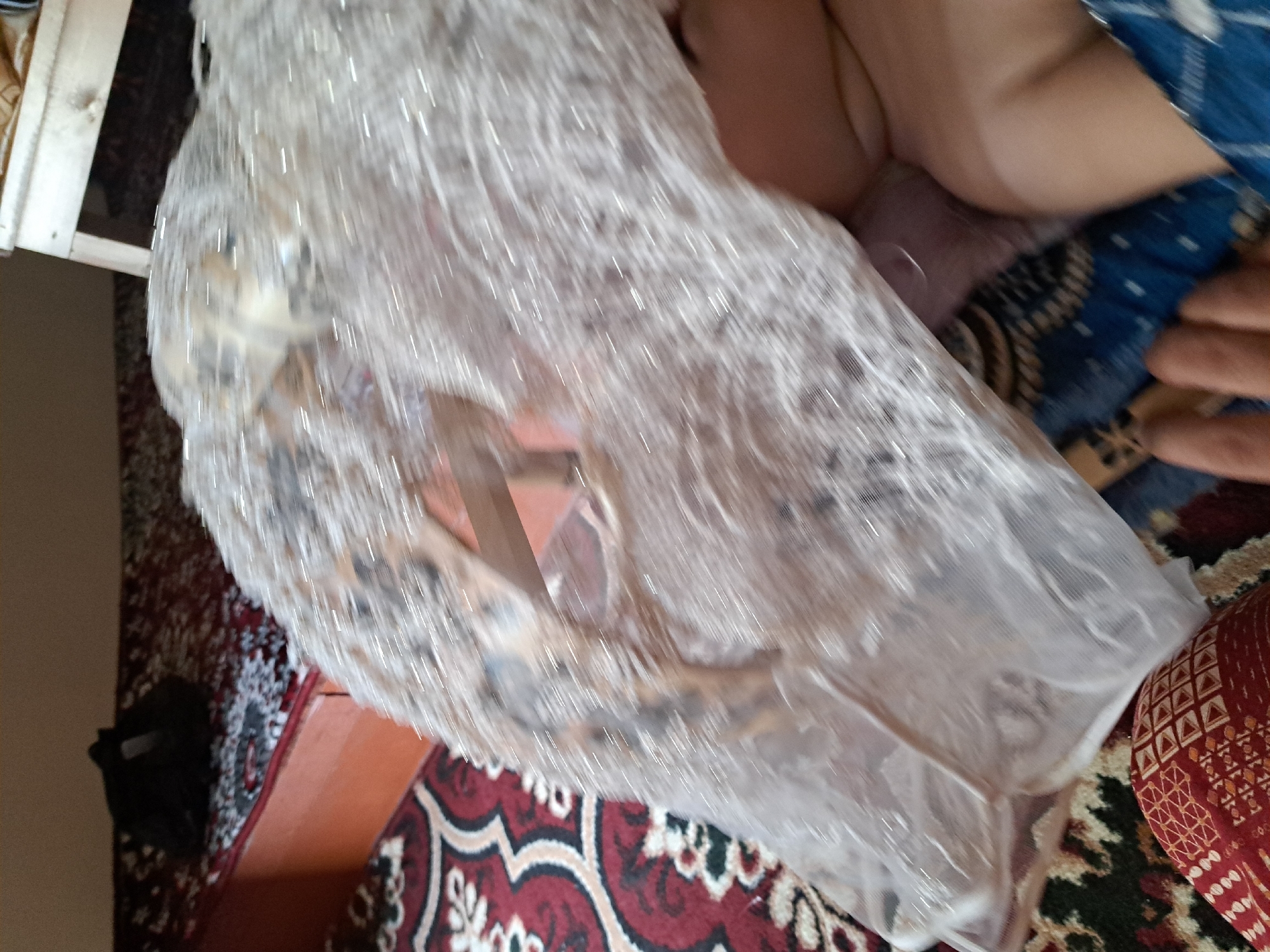 An overhead, slightly blurred medium shot depicts a section of an interior room. In the foreground, a large piece of white, sparkly fabric, possibly a dress or veil, is draped over a person's leg and body, obscuring much of the lower half of the image. The fabric has a sheer, net-like texture with reflective embellishments. To the left, a portion of a red and white patterned carpet is visible, with a dark red border and an intricate floral design. Behind the fabric and to the left, a wooden-framed bed or bench with a red and white patterned mattress is partially visible. On the mattress, a black object, possibly a bag or clothing, rests. In the background, a light-colored wooden stool or table is present. The walls appear to be a neutral, light brown color. The scene suggests a domestic setting, possibly a bedroom or living area in Qarnon, Uzbekistan.