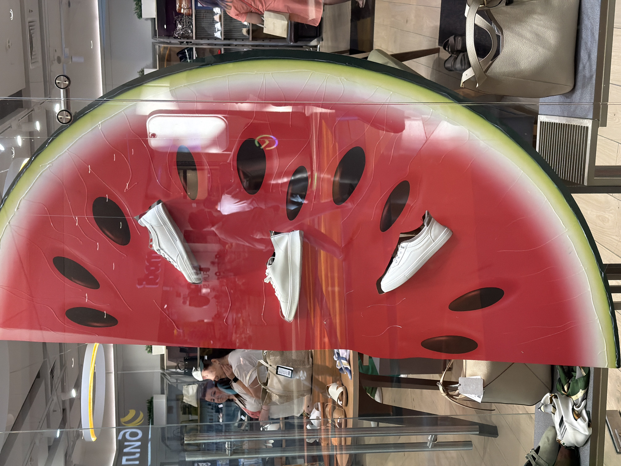 This is a full shot of a store window display in Khimki, Russia, featuring a large, stylized slice of watermelon as the main decorative element. Several pairs of white women's sneakers are showcased on the watermelon design, attached to a red surface. To the left of the watermelon display, behind a glass barrier, a retail environment is visible. Inside, two women are browsing. One woman, wearing a white top, is looking towards the left. The other woman, also in a white top, is facing forward and looking down. A tan handbag is displayed on a wooden table in front of them, along with a pair of sandals. In the foreground on the left, another pair of shoes and a folded scarf are visible on a grey surface. To the right of the watermelon display, a tan handbag is placed on a shelf. The environment appears to be a shoe and accessory store.