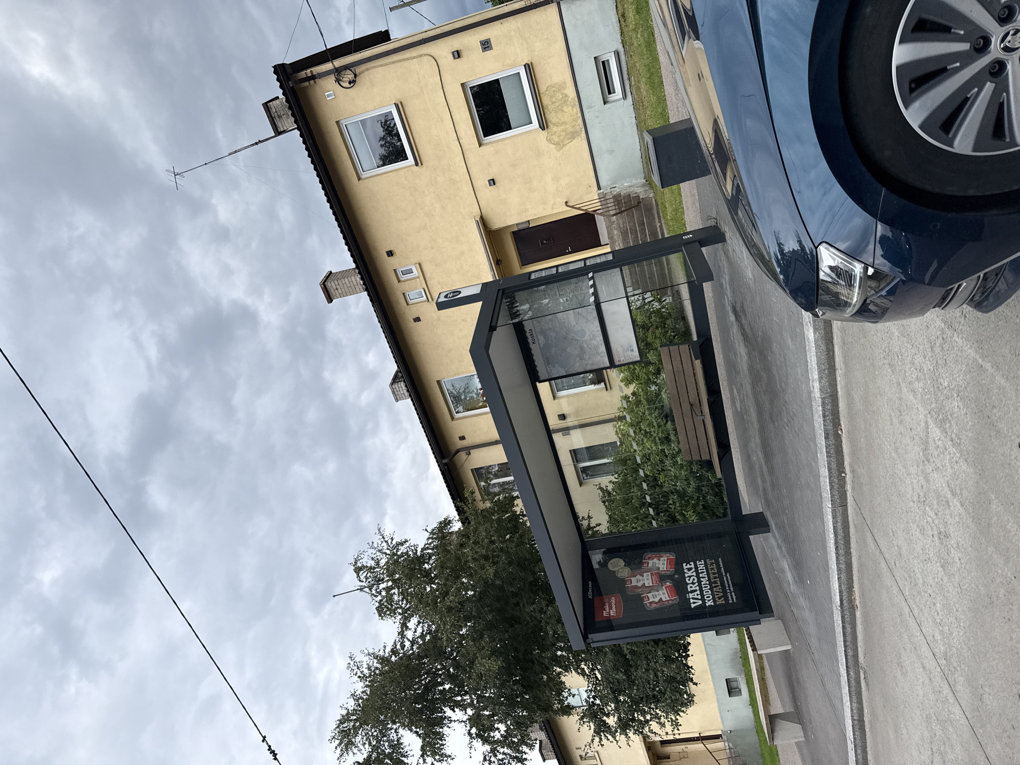 The image depicts a bus stop shelter in Tallinn, Estonia. The shelter has a dark frame and glass panels, with advertisements displayed inside. One advertisement features the text "VÄRSKE KODUMAISE KVALITEET" which translates to "FRESH HOMELAND QUALITY". A dark blue car is partially visible in the foreground, angled towards the right side of the frame. Behind the bus stop, a two-story yellow building with a dark roof and several windows is visible. A large tree with dense foliage stands to the left of the building and bus stop. The sky is overcast with grey clouds. The ground is paved with concrete and asphalt. The scene appears to be an urban street or residential area.