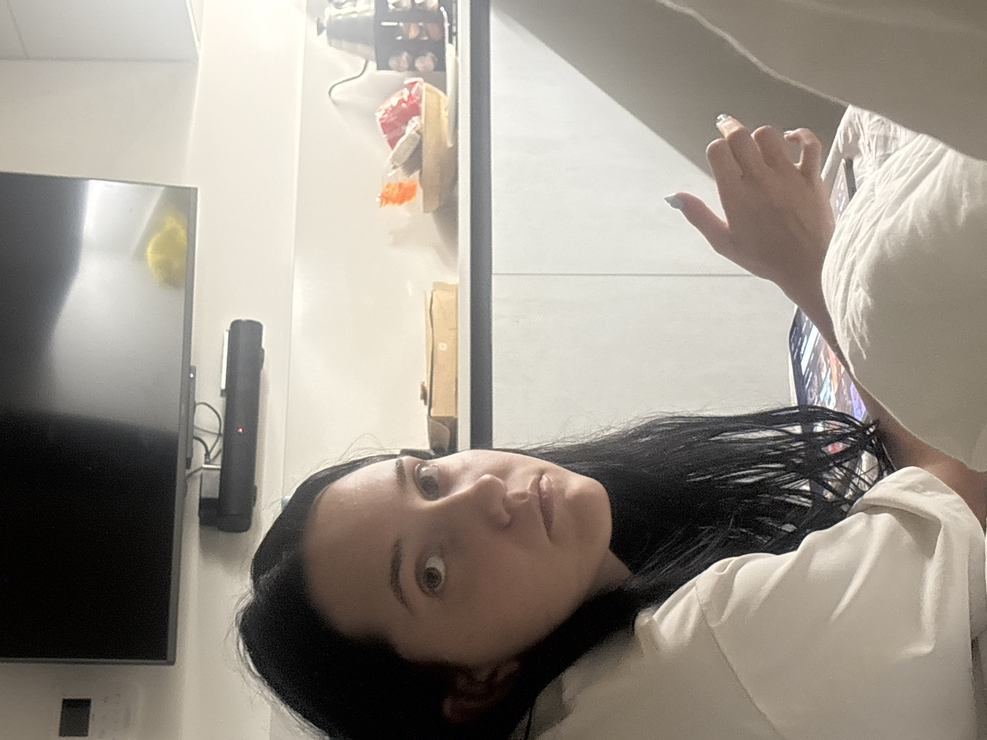 The image shows a young woman with long, dark hair looking towards the camera with a neutral expression. She is wearing a white shirt and appears to be indoors, possibly in a kitchen or living area. In the background, a flat-screen television is mounted on the wall above a soundbar. Further back, a countertop is visible with various items, including a bowl containing food or snacks, a teapot, and what appears to be a bag of chips. The woman's right hand is resting on a white surface, and a tablet or similar electronic device is partially visible beneath her arm. The lighting suggests it is daytime. The scene is not discernibly located in Cupra Marittima, Italy, or any specific city.