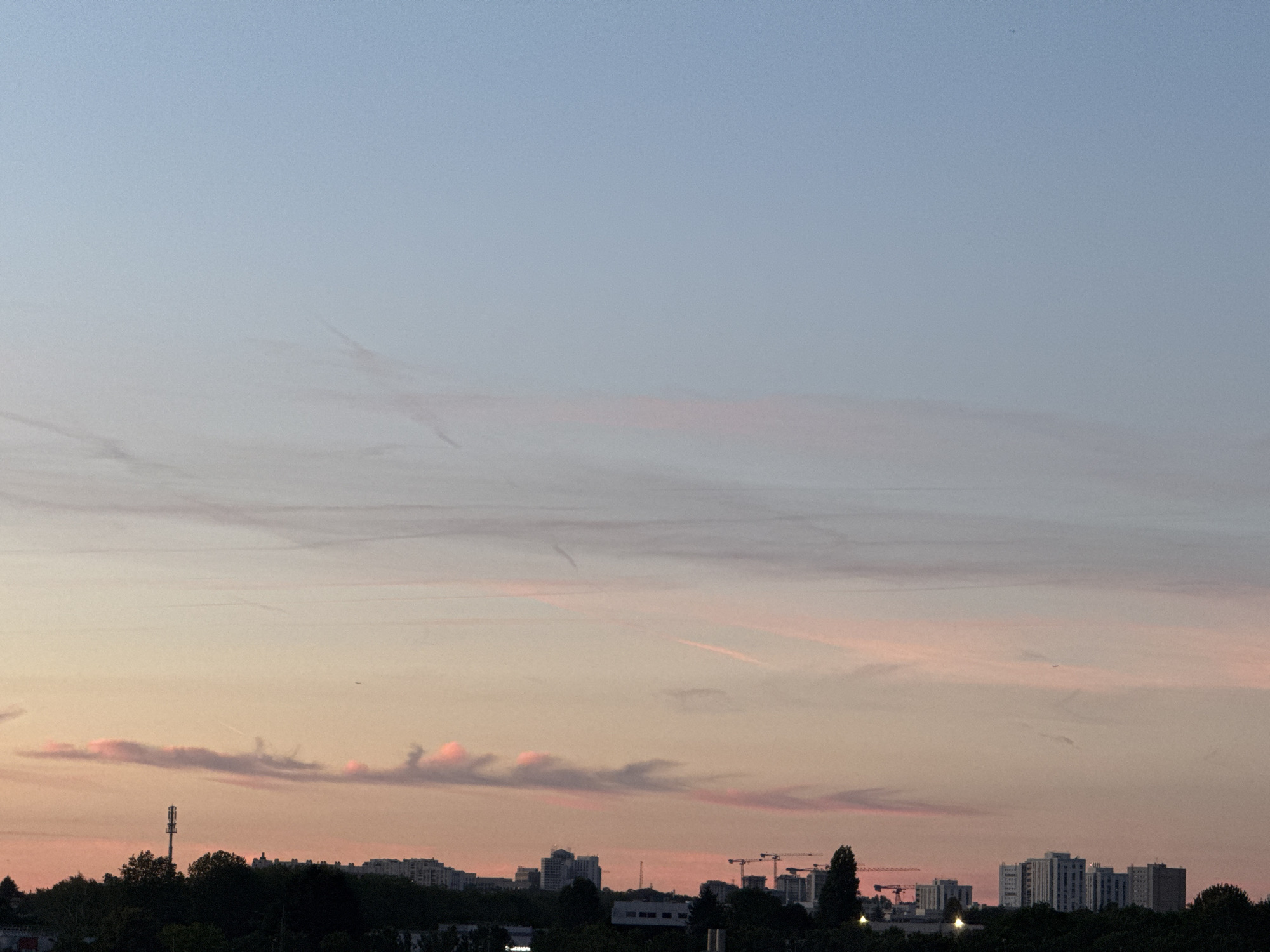 The image shows a cityscape at dusk. The sky is a gradient of light blue to pale pink, with thin, wispy clouds. Several contrails are visible in the sky, indicating air traffic. In the foreground, silhouetted against the sky, is a dense line of trees, interspersed with buildings. A cellular tower stands prominently on the left side of the frame. Further into the cityscape, apartment buildings and construction cranes are visible. The overall impression is of an urban environment transitioning from day to night.